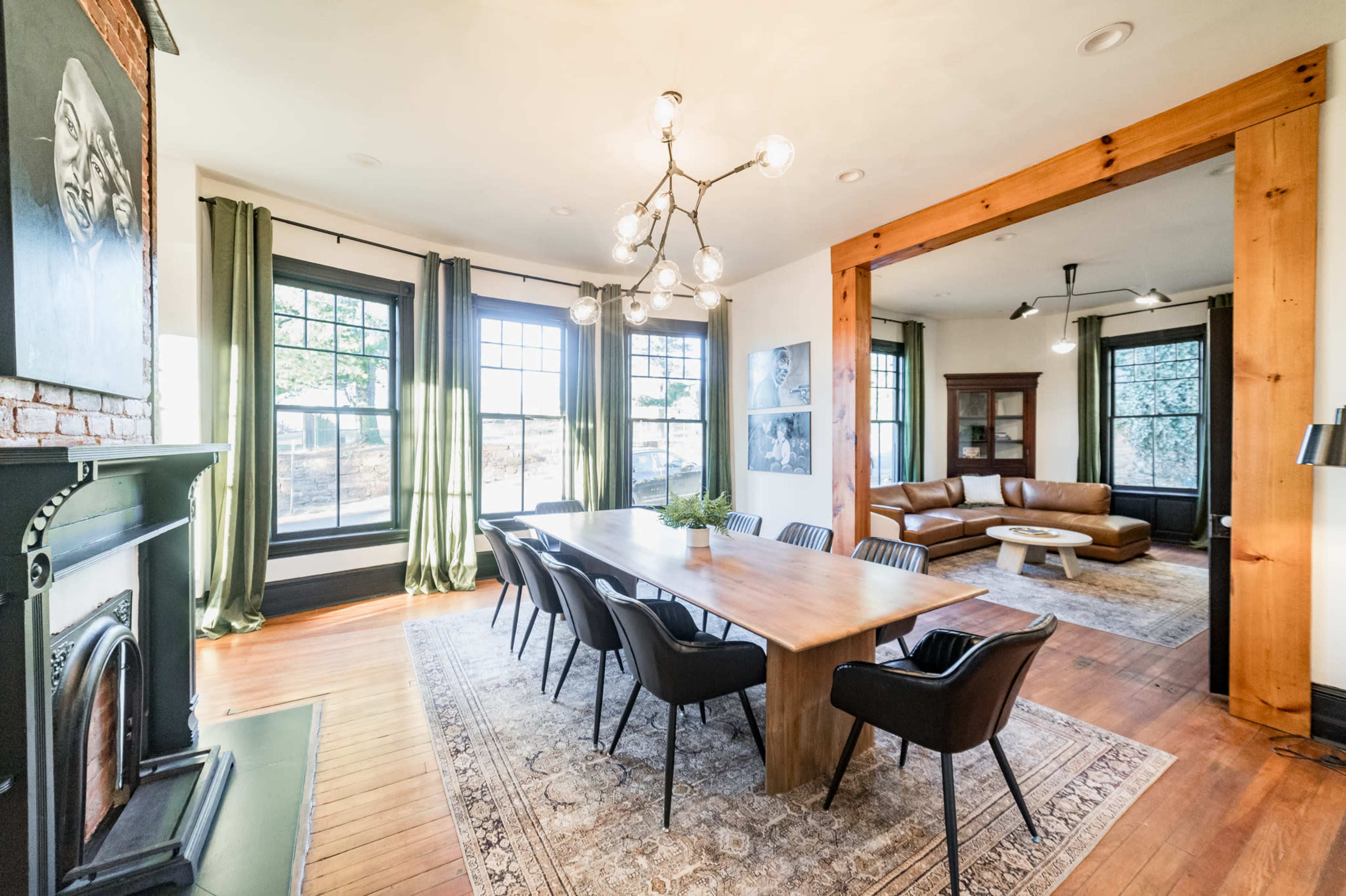 A dining area with a wooden table and black chairs is adjacent to a living space featuring a brown leather sofa and large windows.