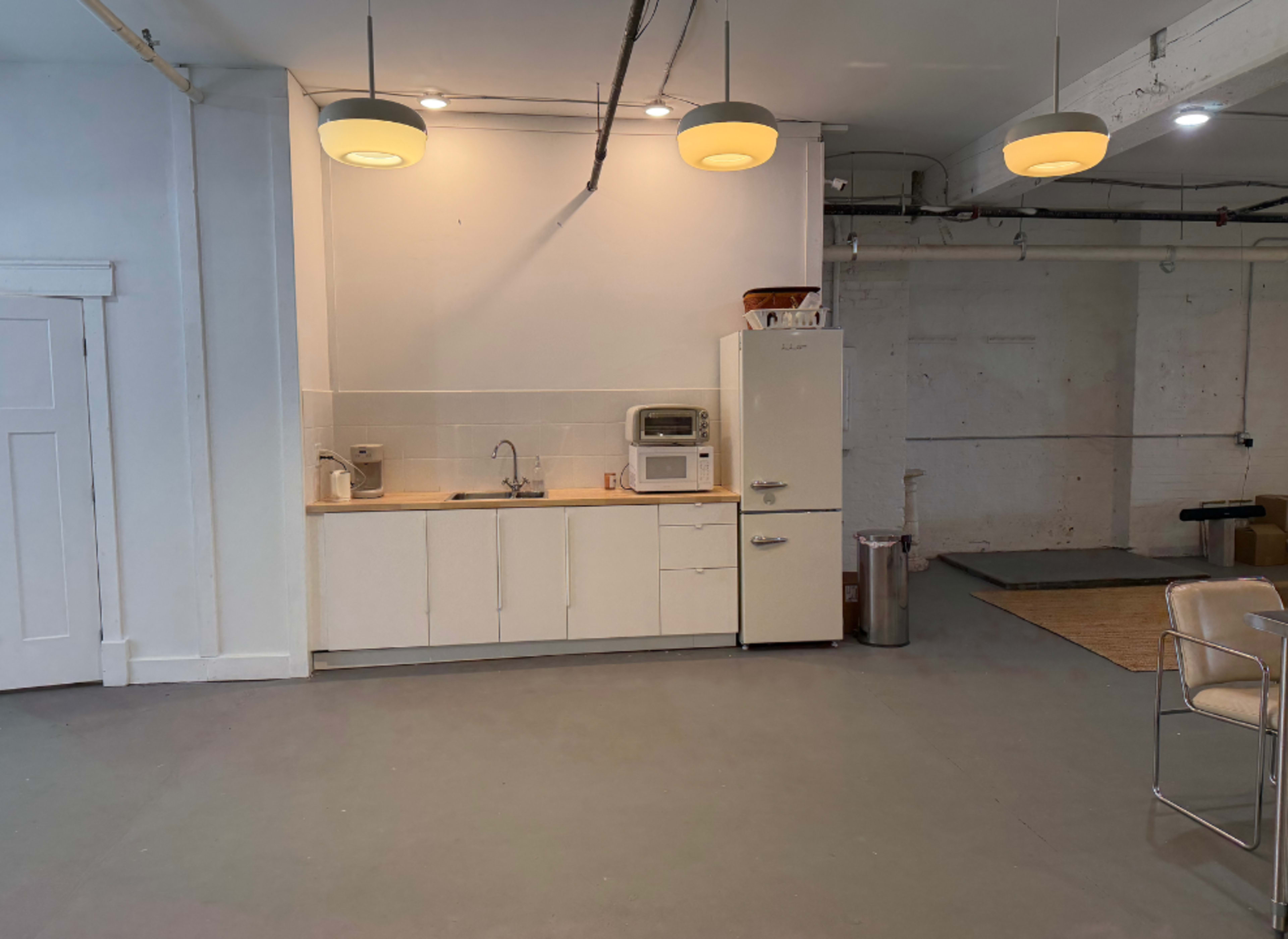 The image shows a spacious interior with a kitchenette consisting of a sink, a microwave, and a refrigerator, along with light fixtures hanging from the ceiling.