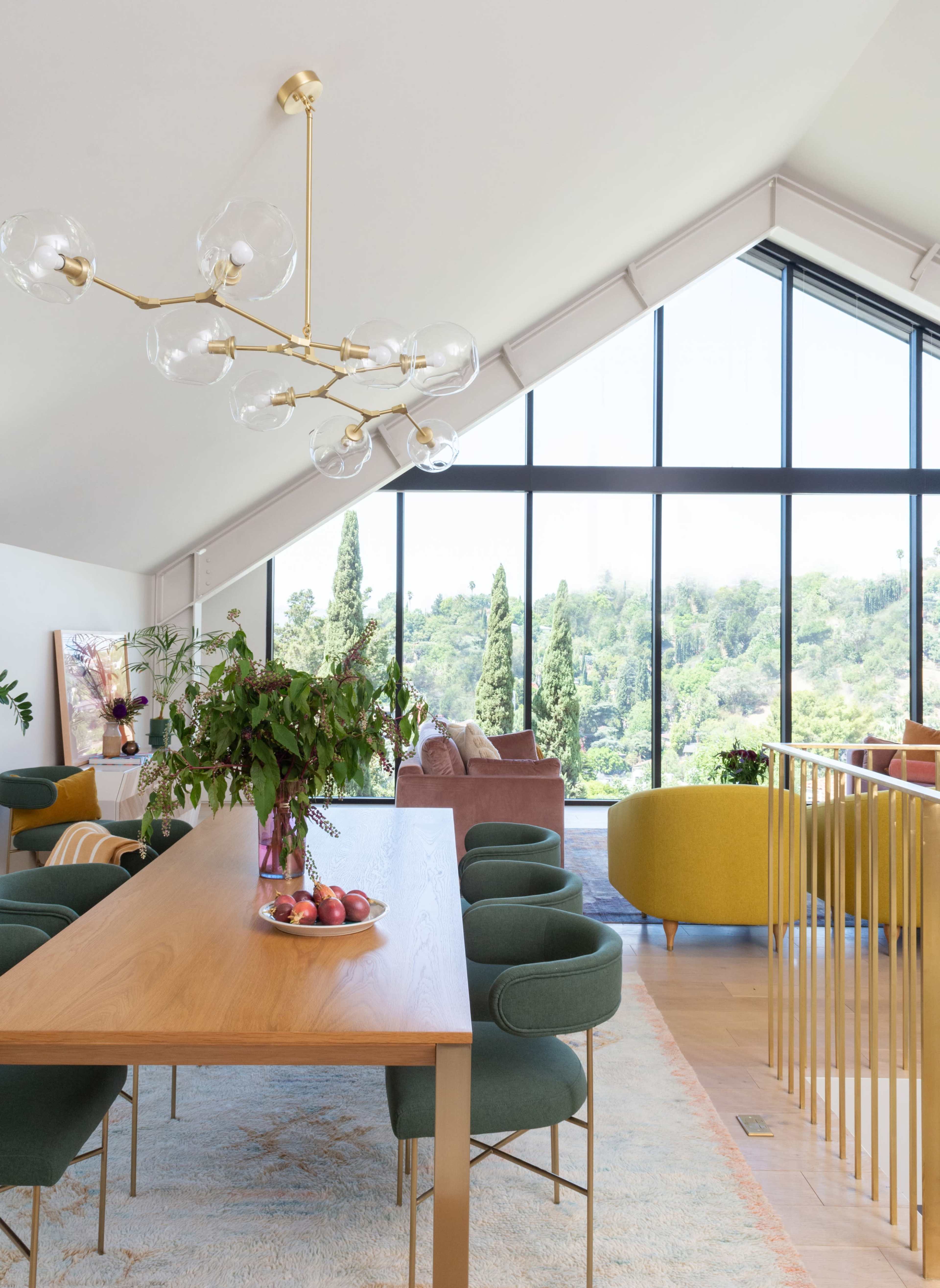 A modern dining area features a wooden table surrounded by green chairs, with large windows offering a view of trees and hills beyond.