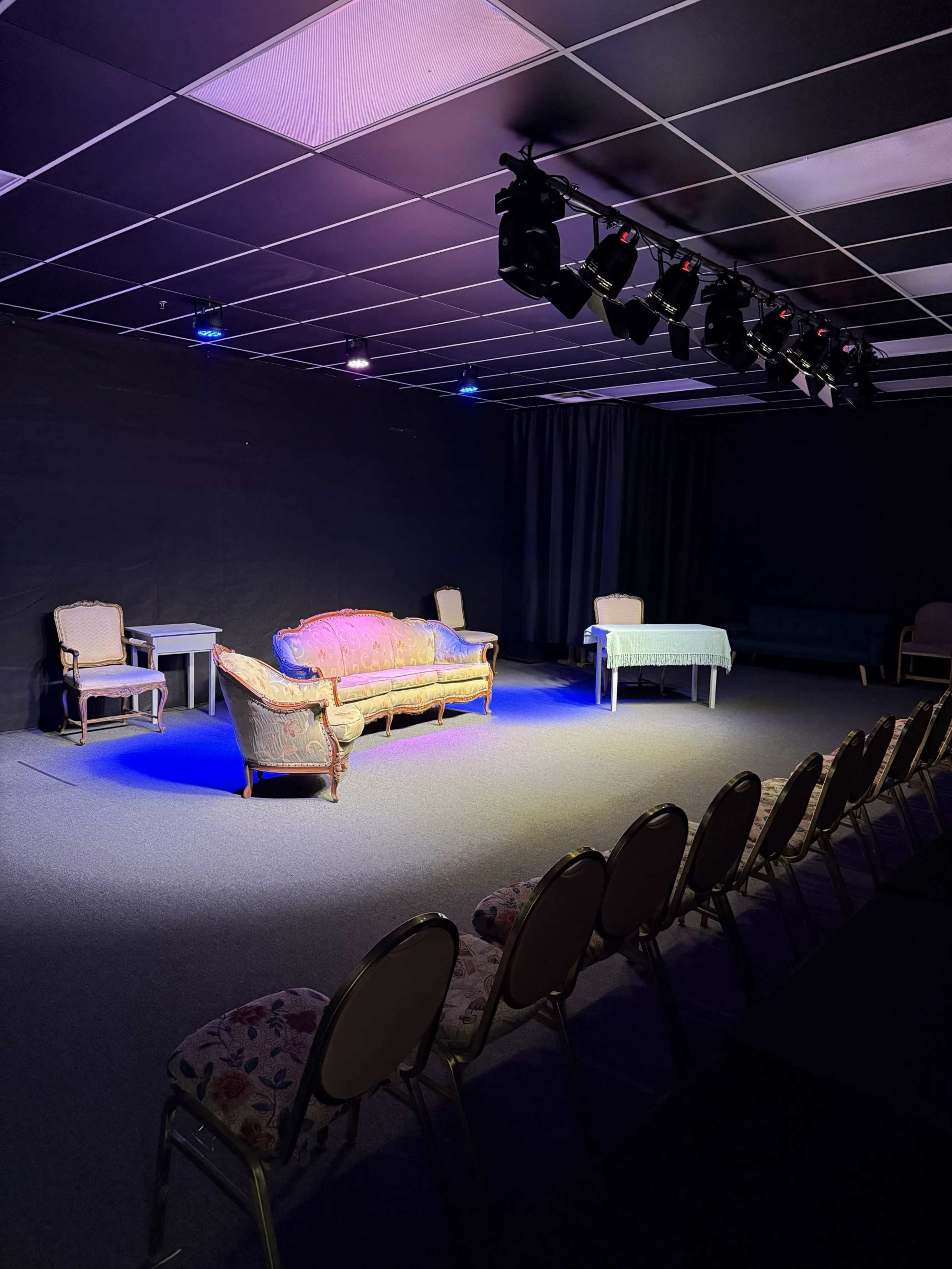 The image shows a small stage set with a colorful sofa, two chairs, a table, and rows of empty chairs arranged in front of it under theatrical lighting.