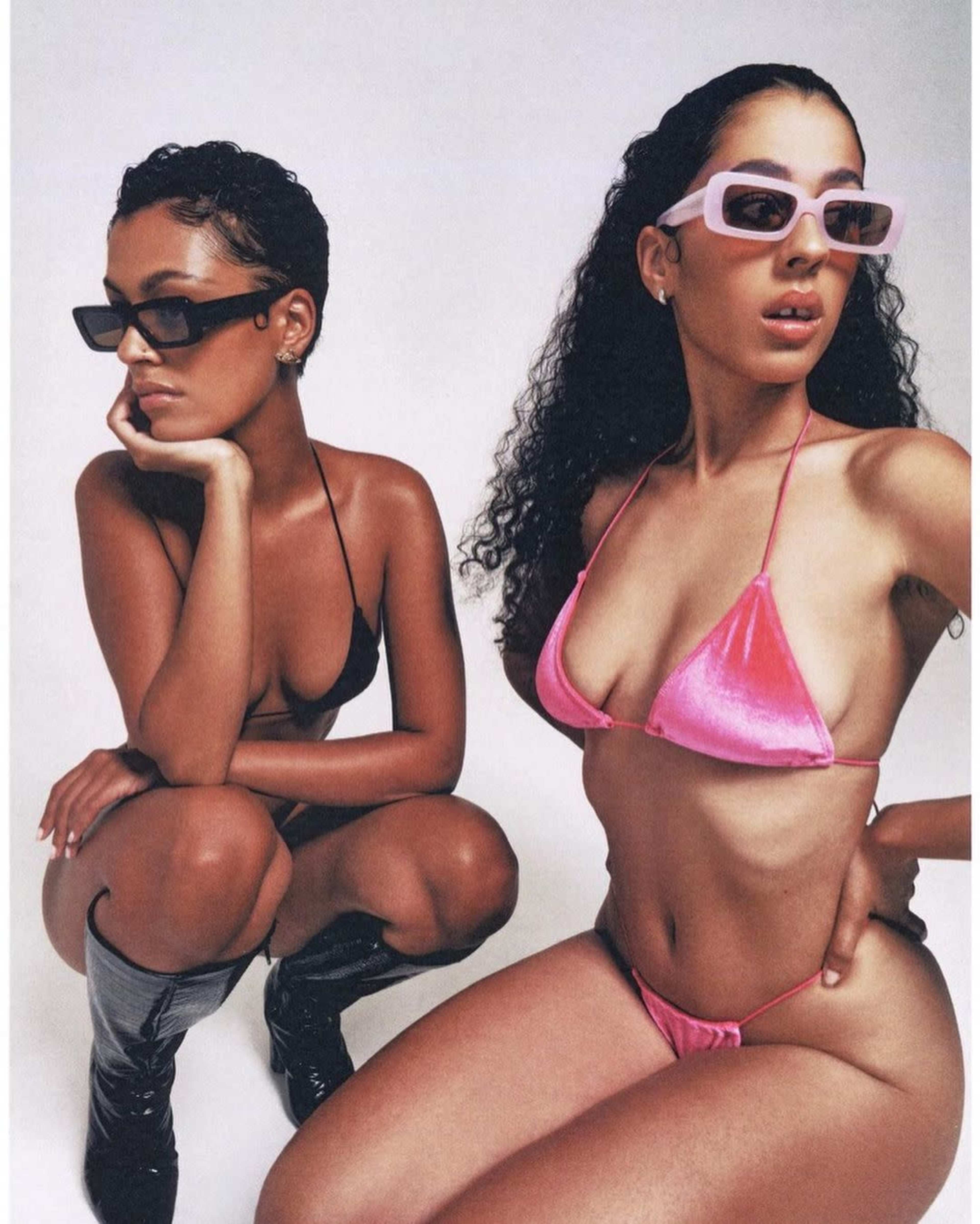 Two women pose in stylish swimwear, one in a black bikini and the other in a pink bikini, both wearing modern sunglasses.