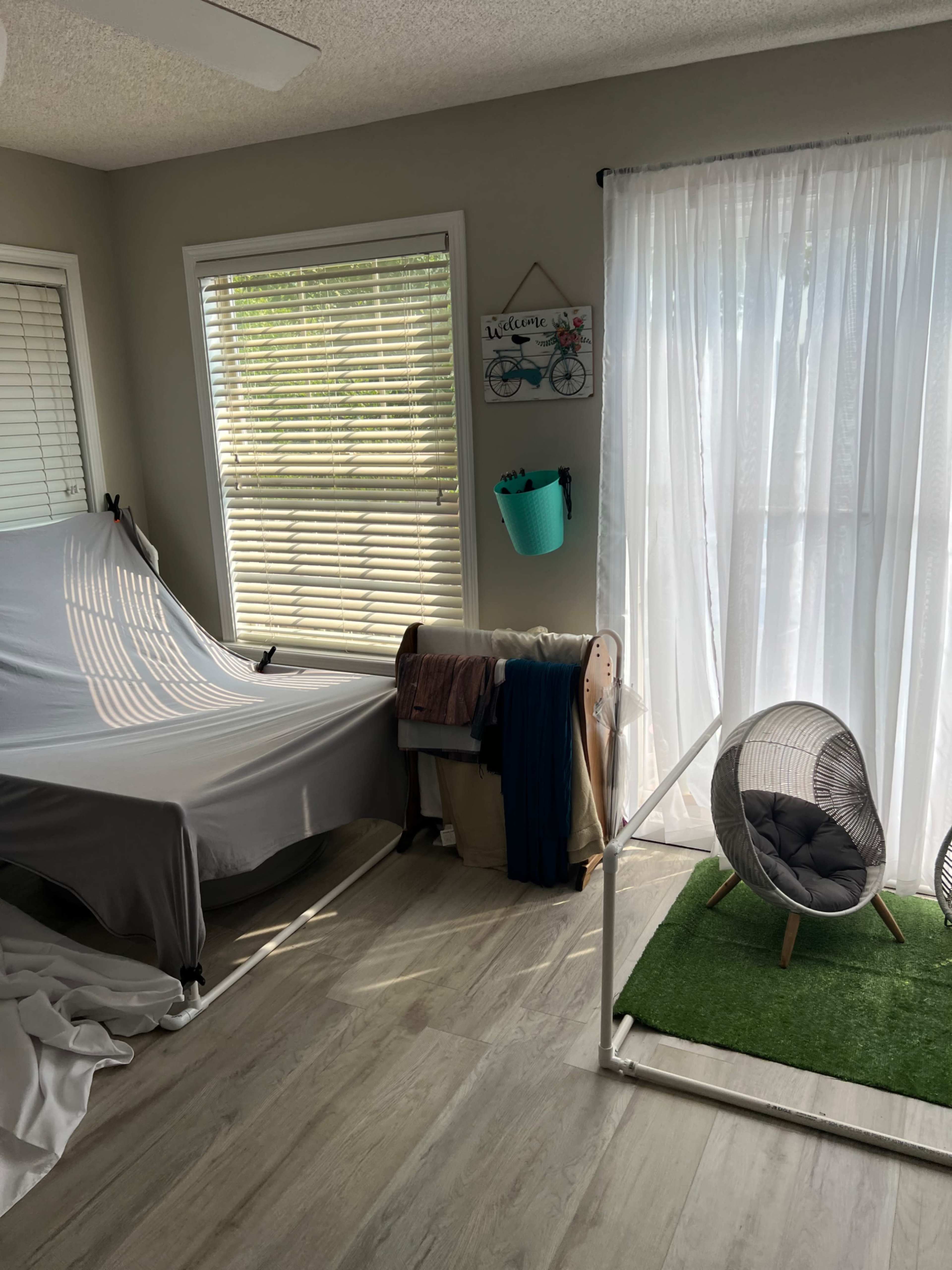 A sunny room with a covered sofa, a folding chair, a decorative wall sign, and sheer curtains letting in light.