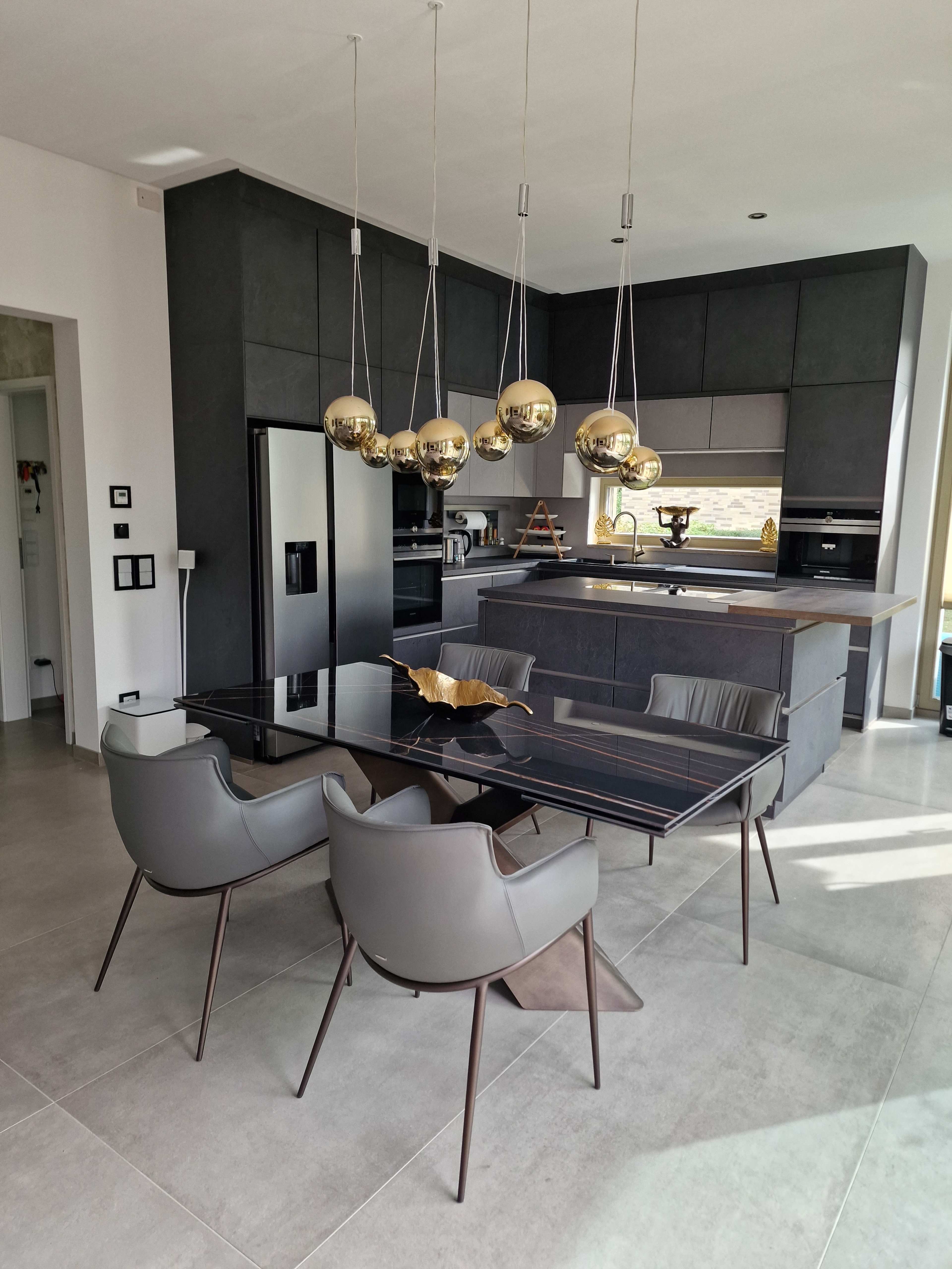 A modern kitchen features a sleek black countertop, metallic pendant lights, and a glass dining table surrounded by stylish chairs.
