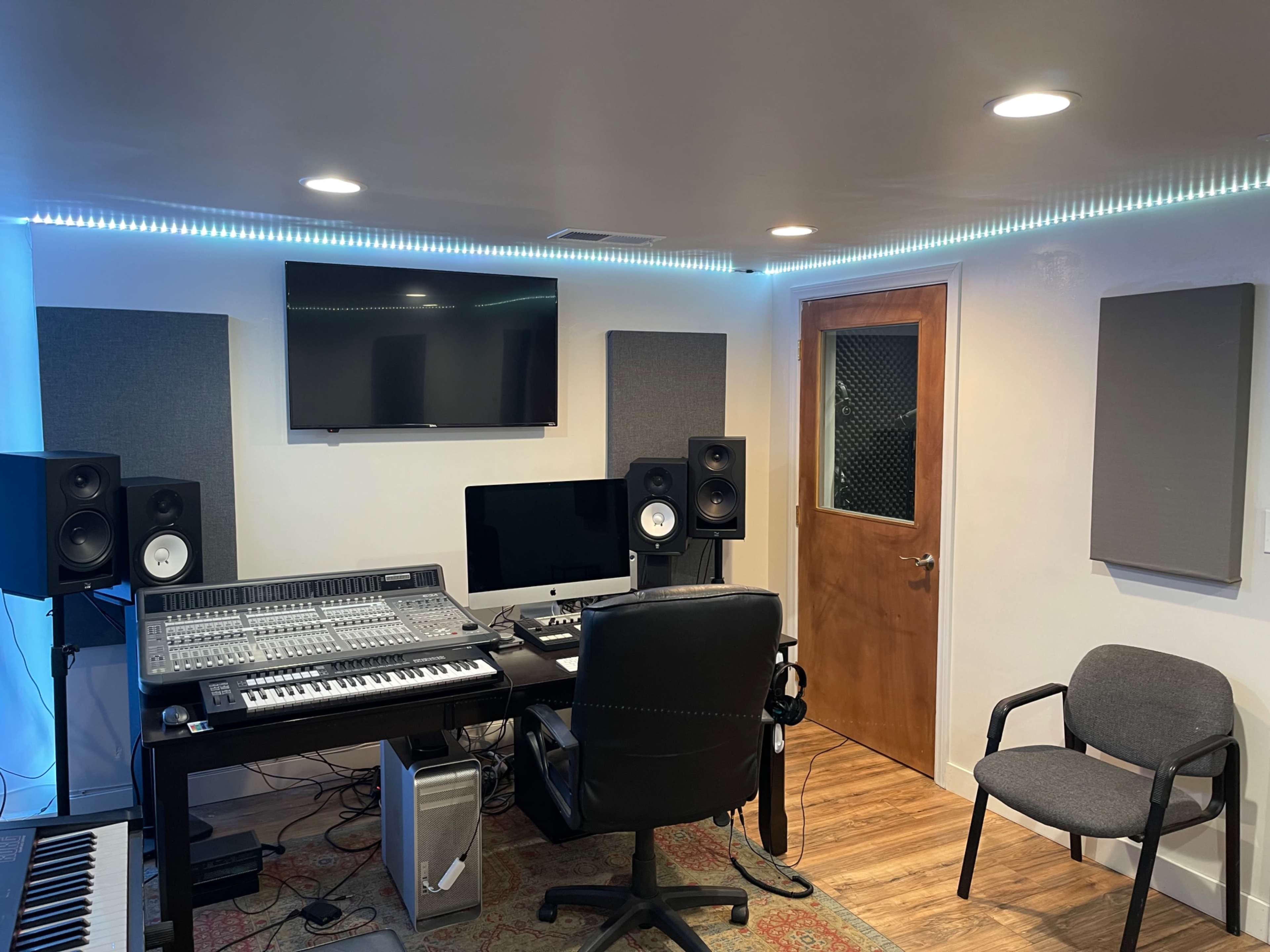 The image shows a modern music studio featuring a mixing console, two speakers, a computer, and soundproofing panels, with a chair and door in the background.