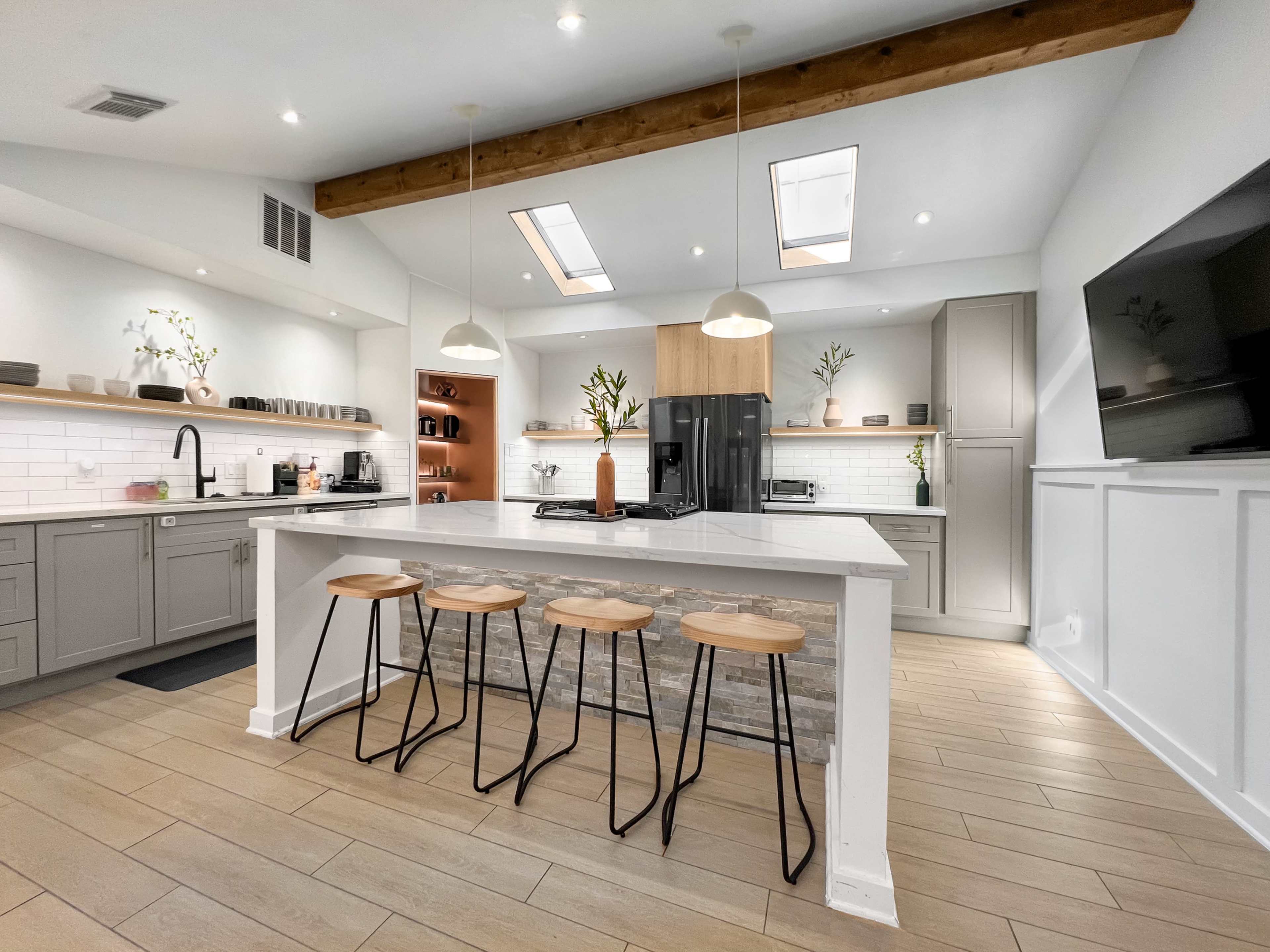 A modern kitchen features an island with four bar stools, gray cabinetry, and a mixture of natural and artificial lighting.