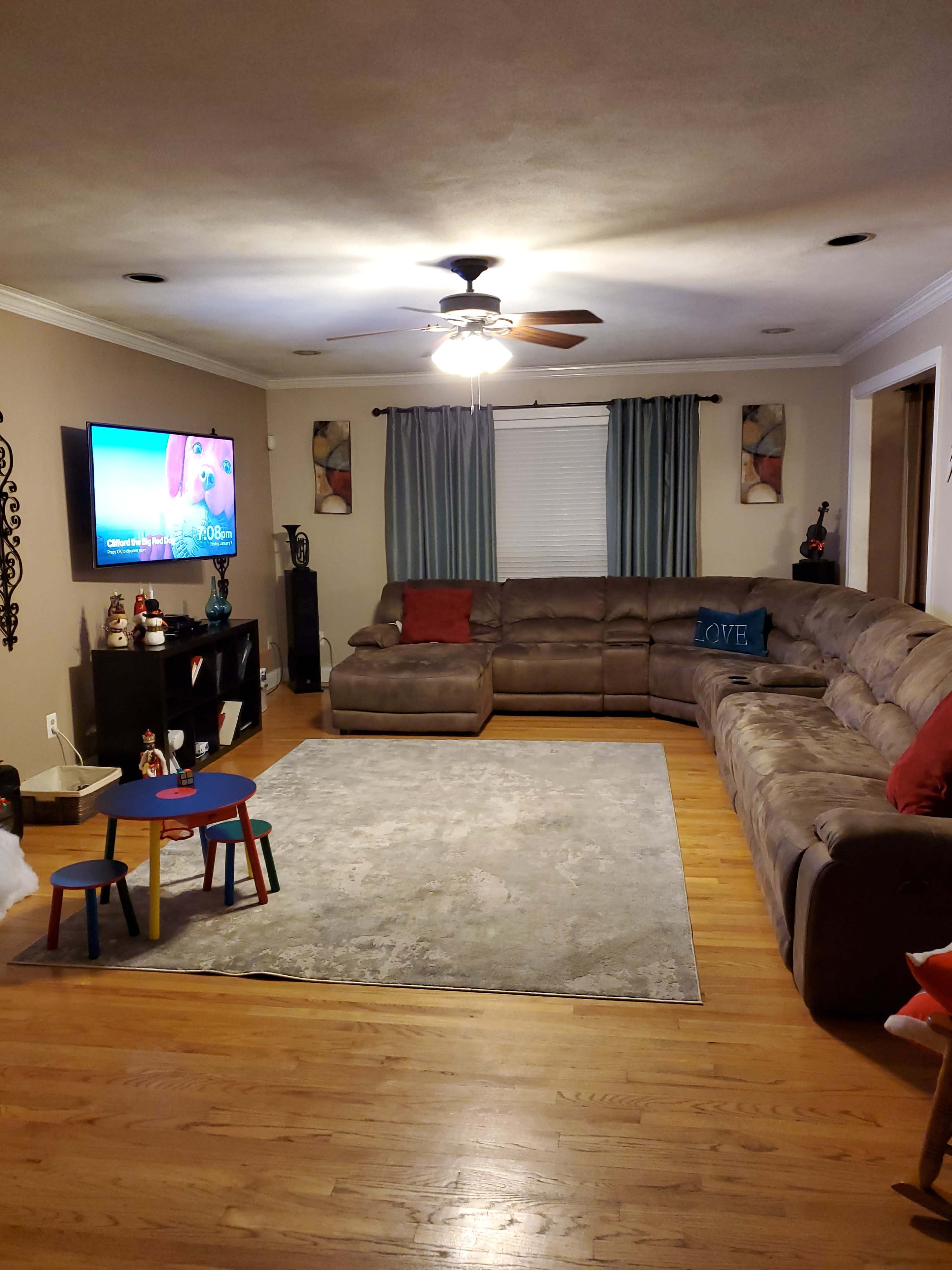 A spacious living room features a large sectional sofa, a coffee table, a television displaying a children's program, and a decorative rug on the wooden floor.