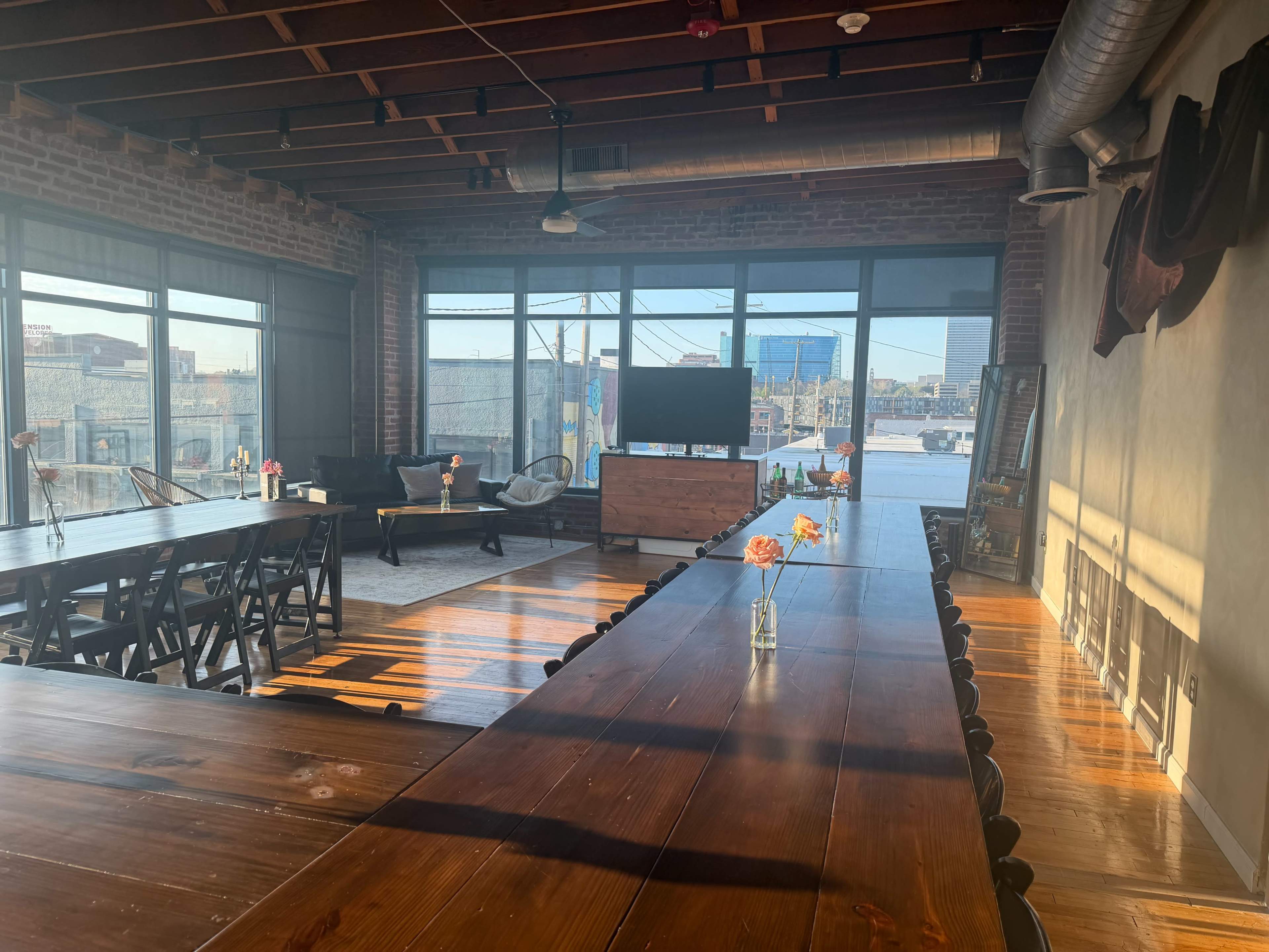 A spacious conference room features large windows with city views, wooden tables, and small flower arrangements on each table.