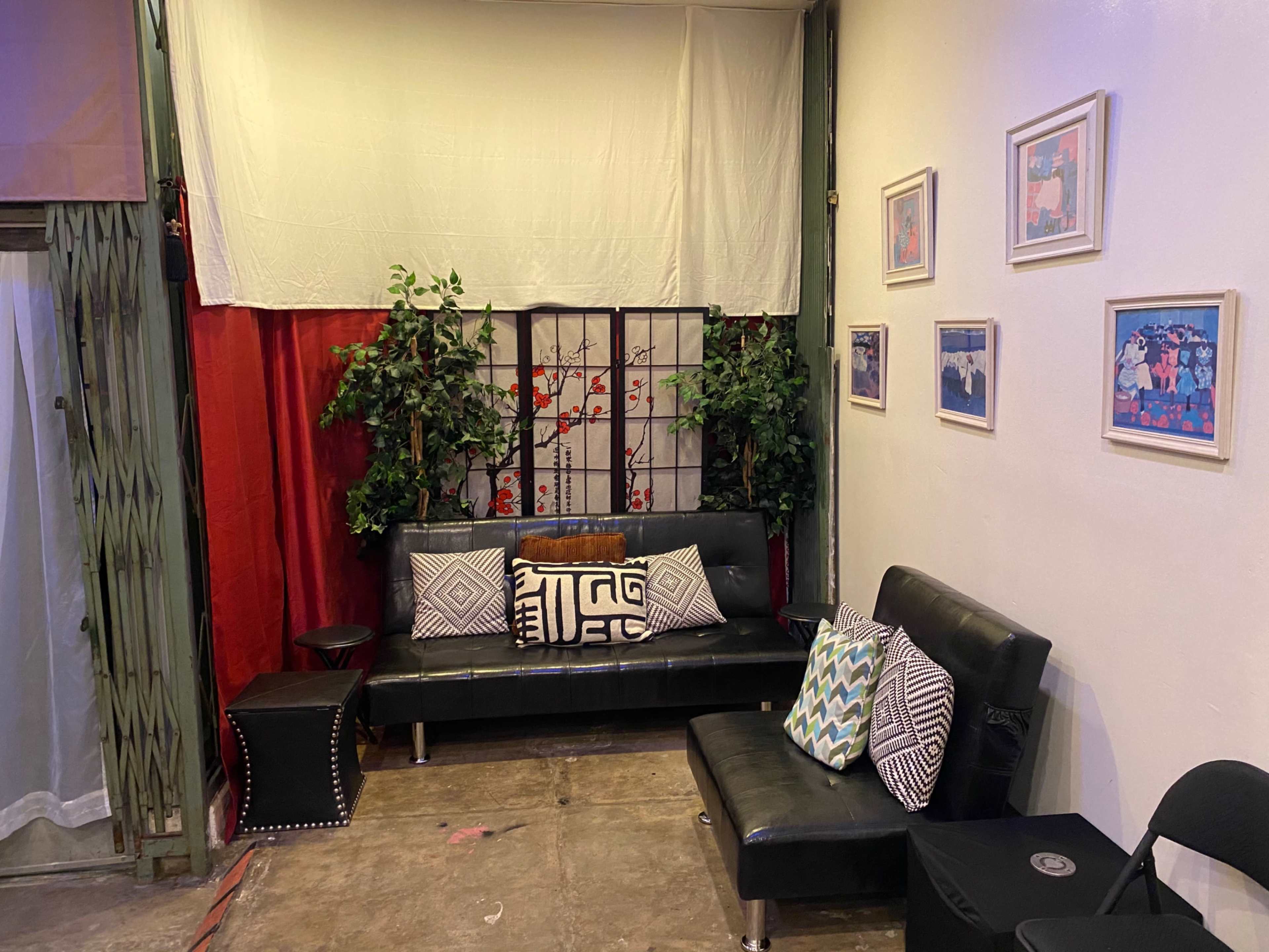 A small, furnished corner with a black leather sofa, decorative pillows, potted plants, and framed artwork on the walls.