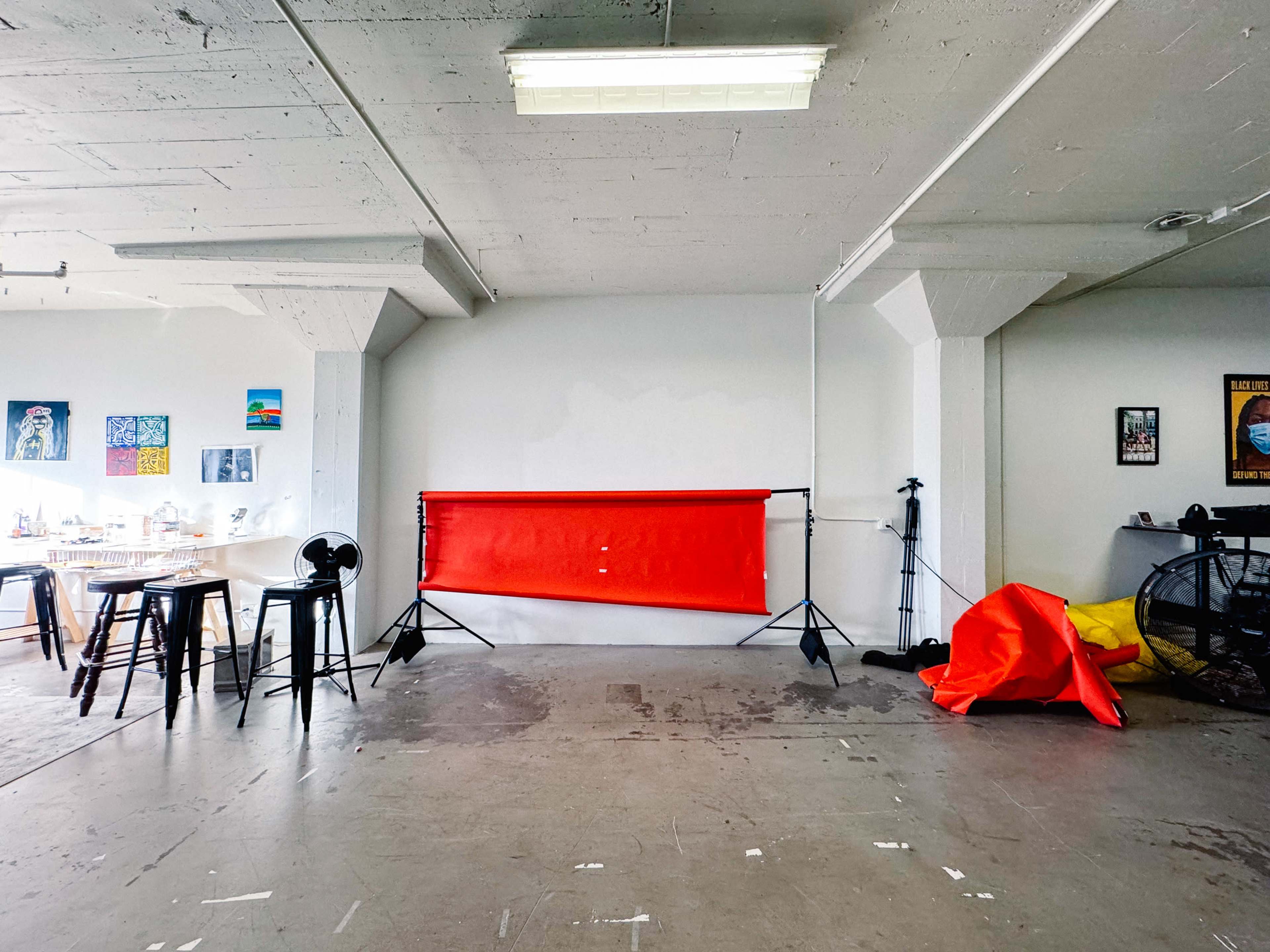 The image shows a spacious room with a red photographic backdrop on stands, a table with chairs, a fan, and a yellow object on the floor.