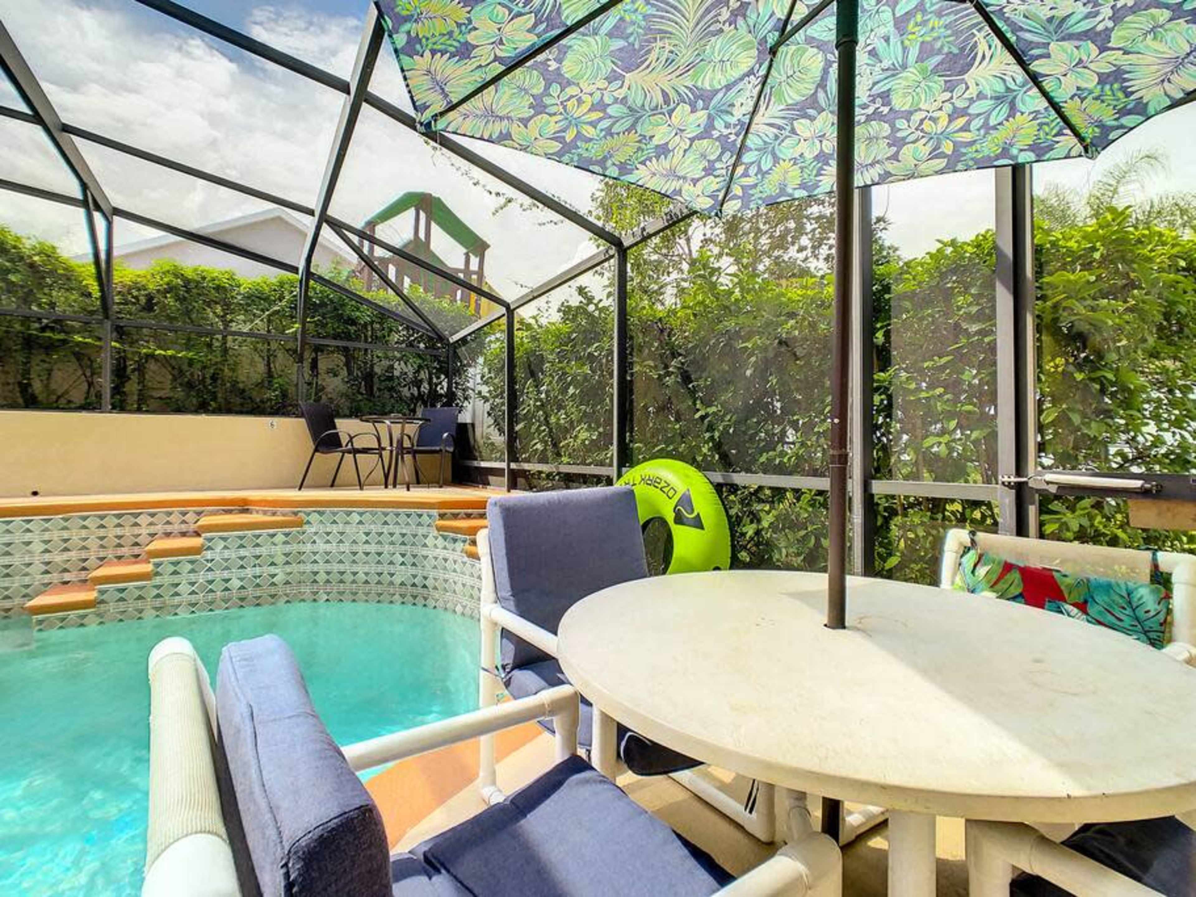 The image shows a screened-in patio with a swimming pool, a round table with chairs underneath an umbrella, and a green inflatable ring leaning against the wall.