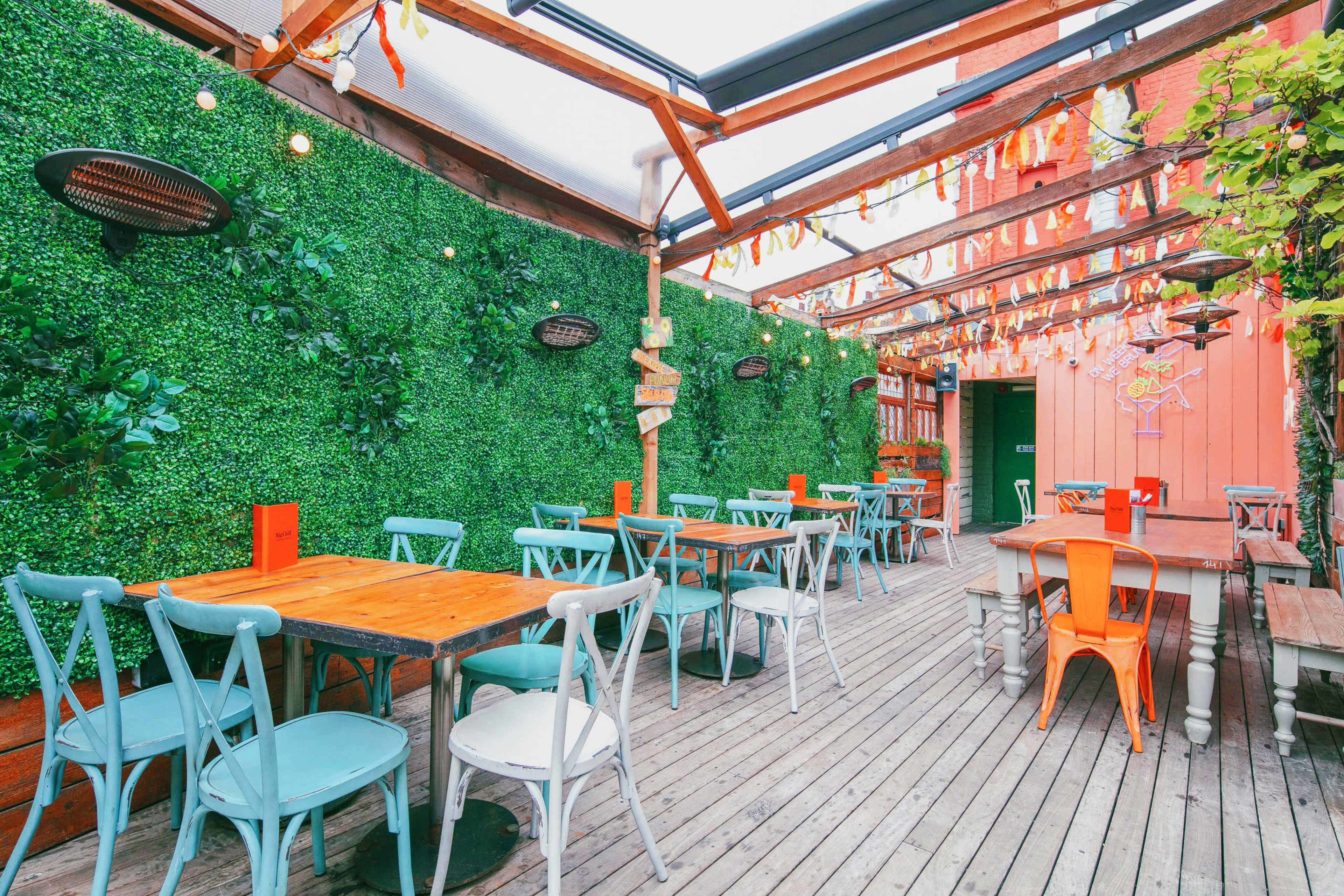 A brightly lit outdoor dining area with wooden tables and colorful chairs, surrounded by greenery and hanging decorations.