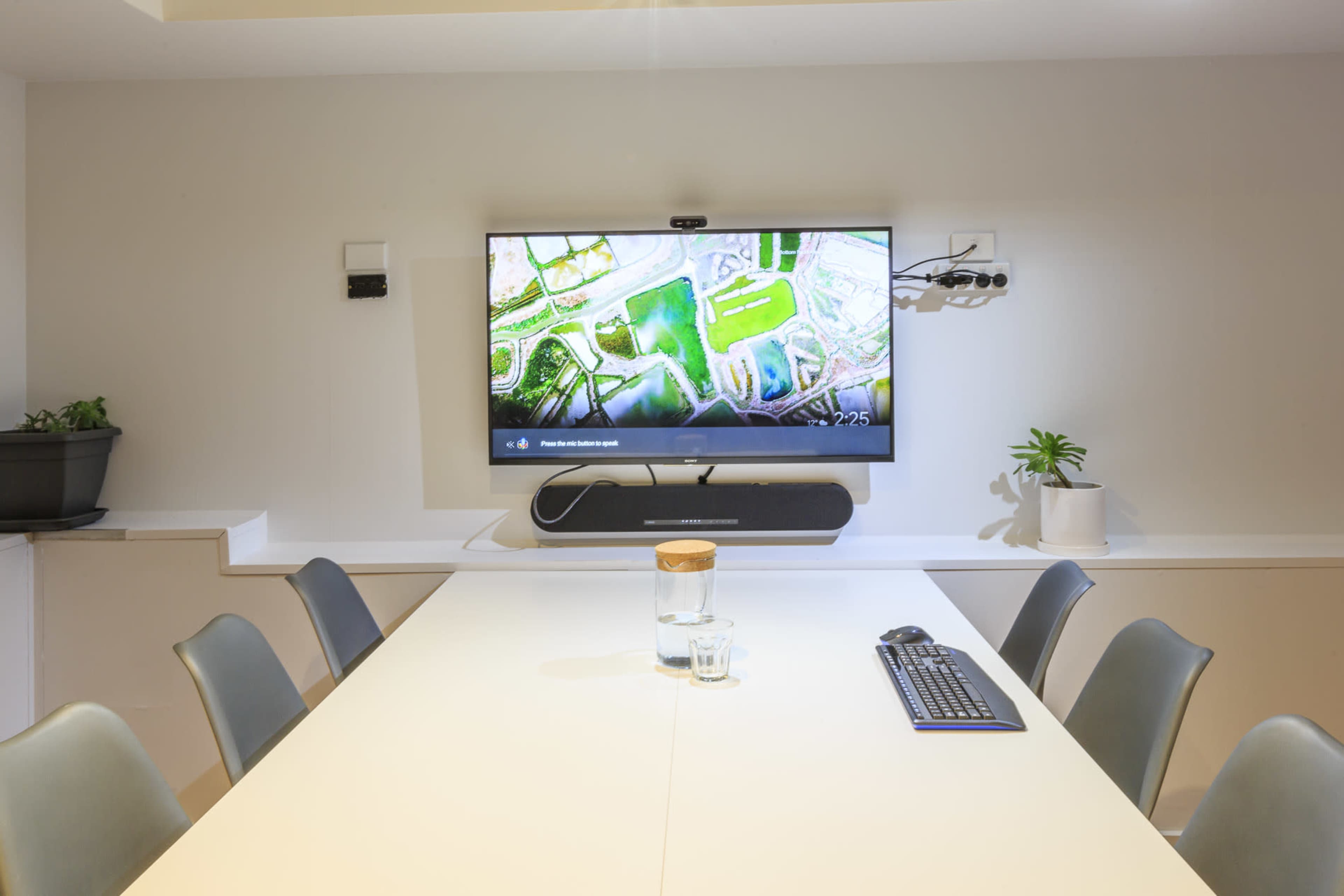 A modern conference room features a large screen displaying a map, with a long table, chairs, a keyboard, a glass of water, and potted plants visible.