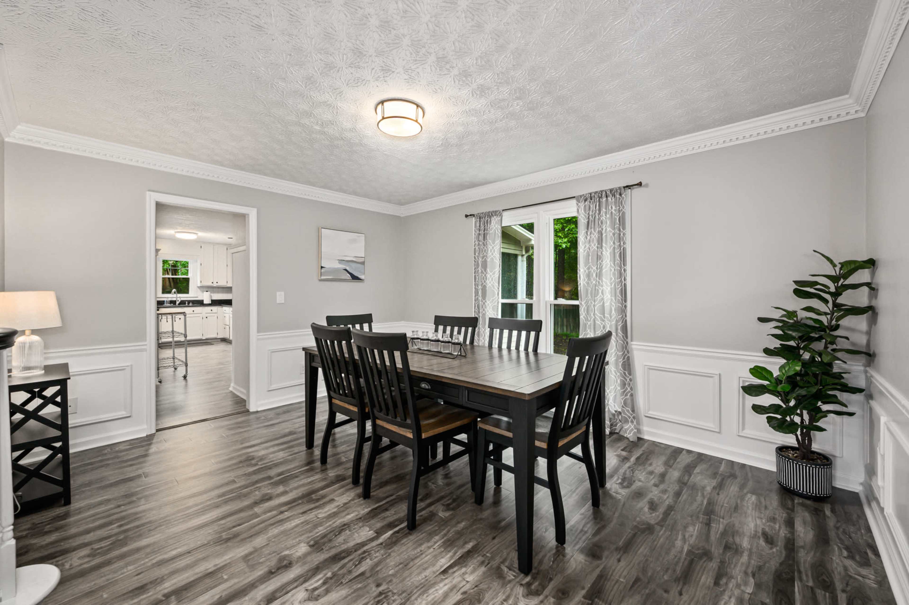 A dining room features a large wooden table surrounded by black chairs, with a window allowing natural light and a view of greenery outside.