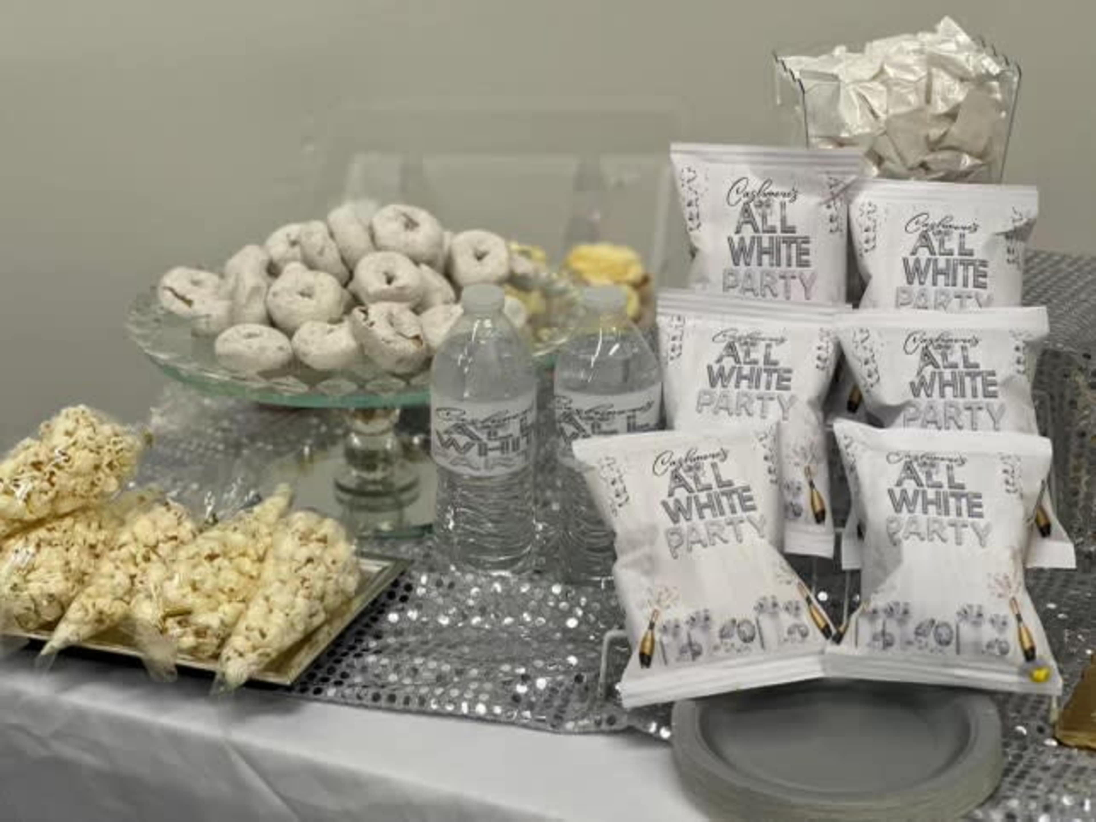 A table is set with various snacks, including donuts, popcorn, and packaged treats, along with bottled water labeled for an "All White Party."