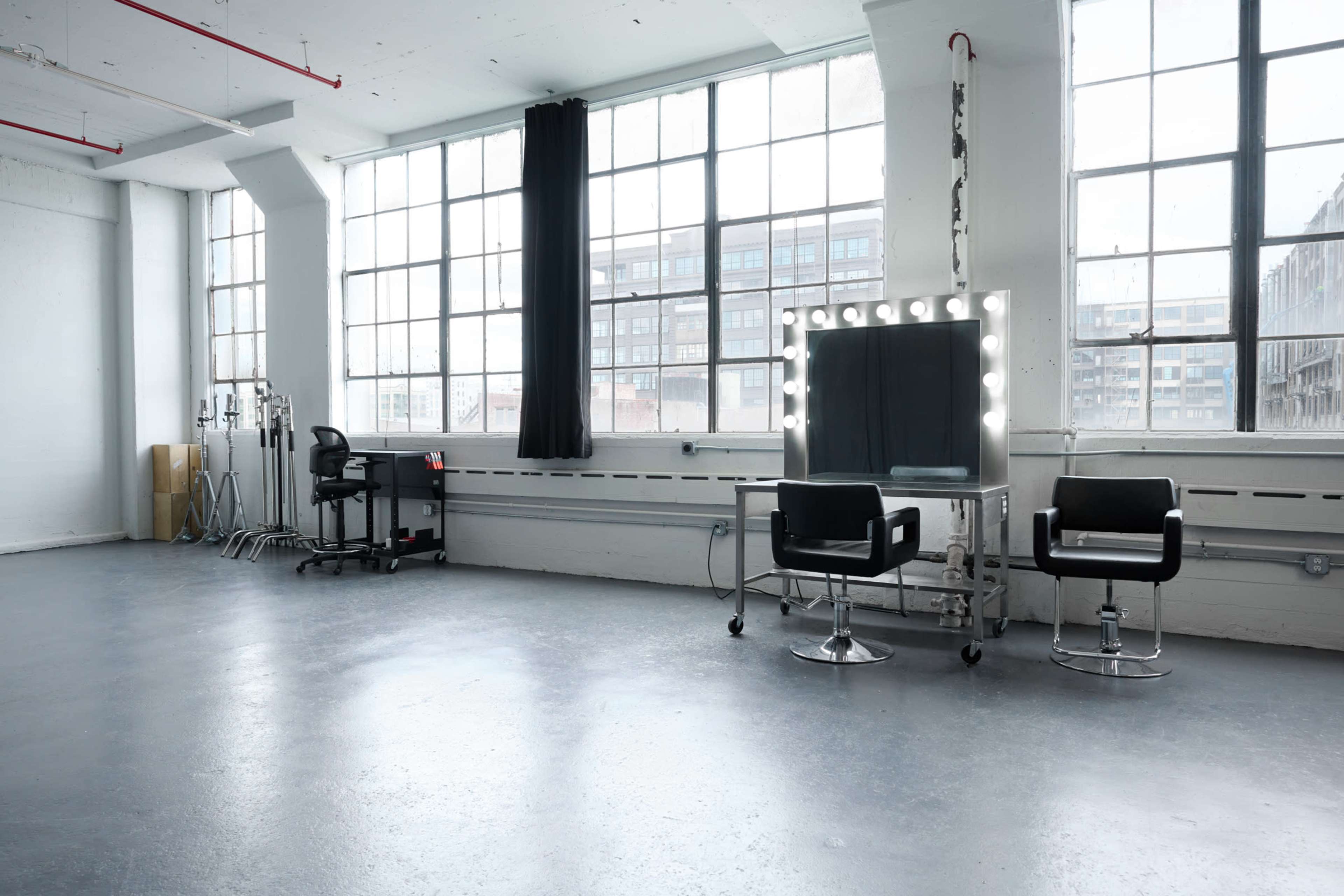 A spacious, well-lit room features large windows, a mirrored vanity with lights, and two black chairs.