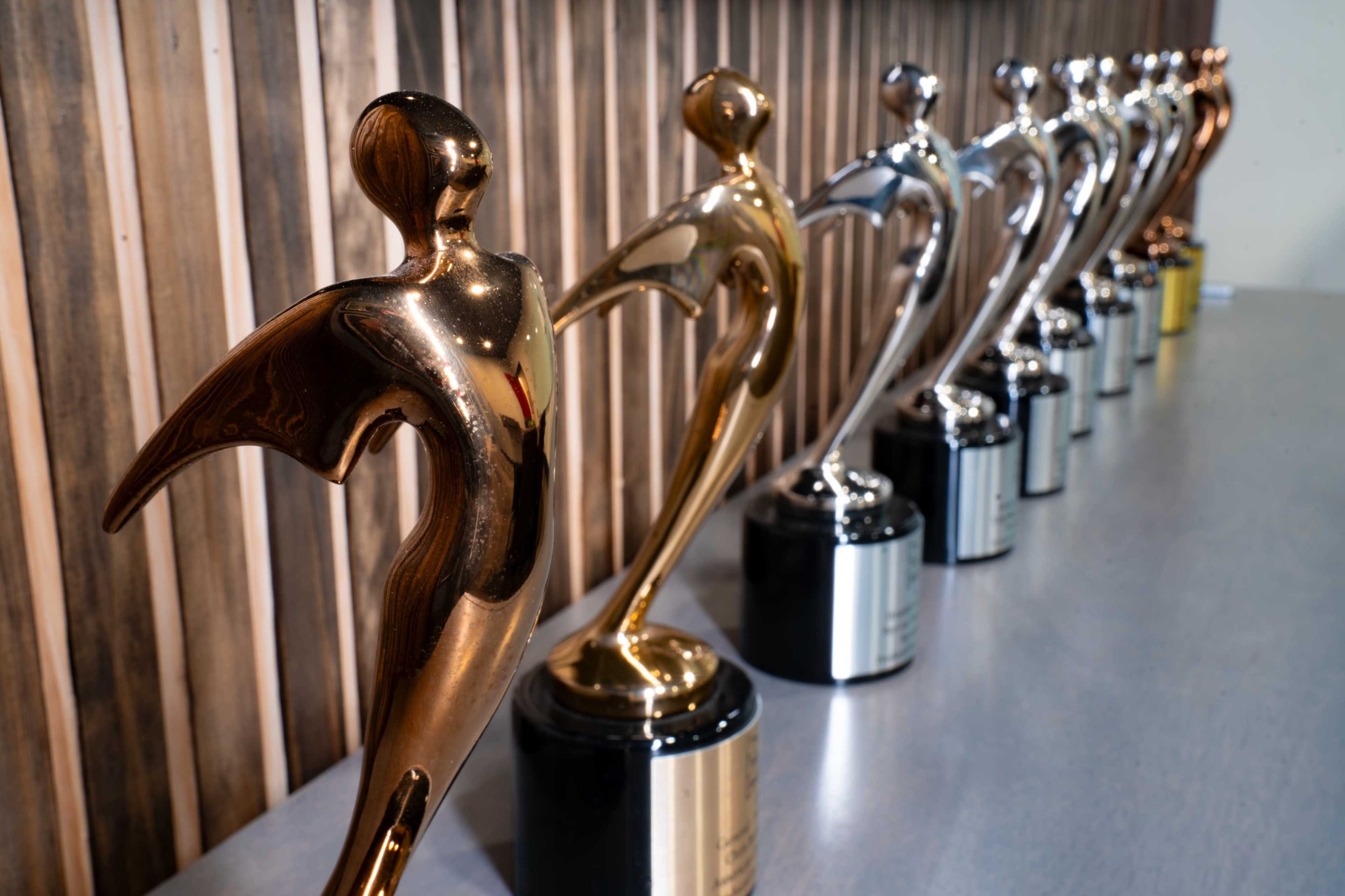 A row of metallic trophies with various finishes stands on a table against a wooden backdrop.
