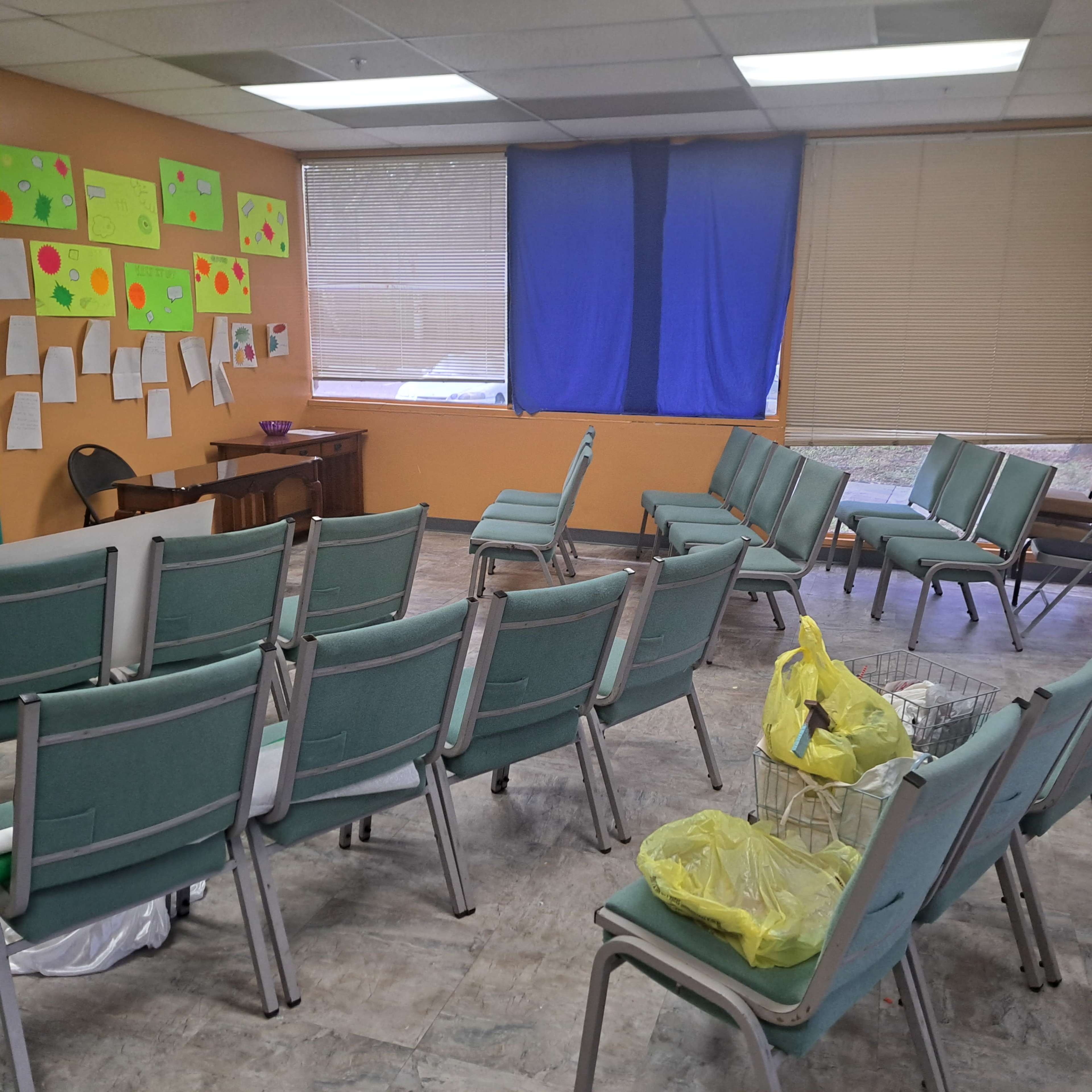 The room features several rows of green chairs arranged for seating, with a blue curtain covering a window and colorful chart paper on the walls.