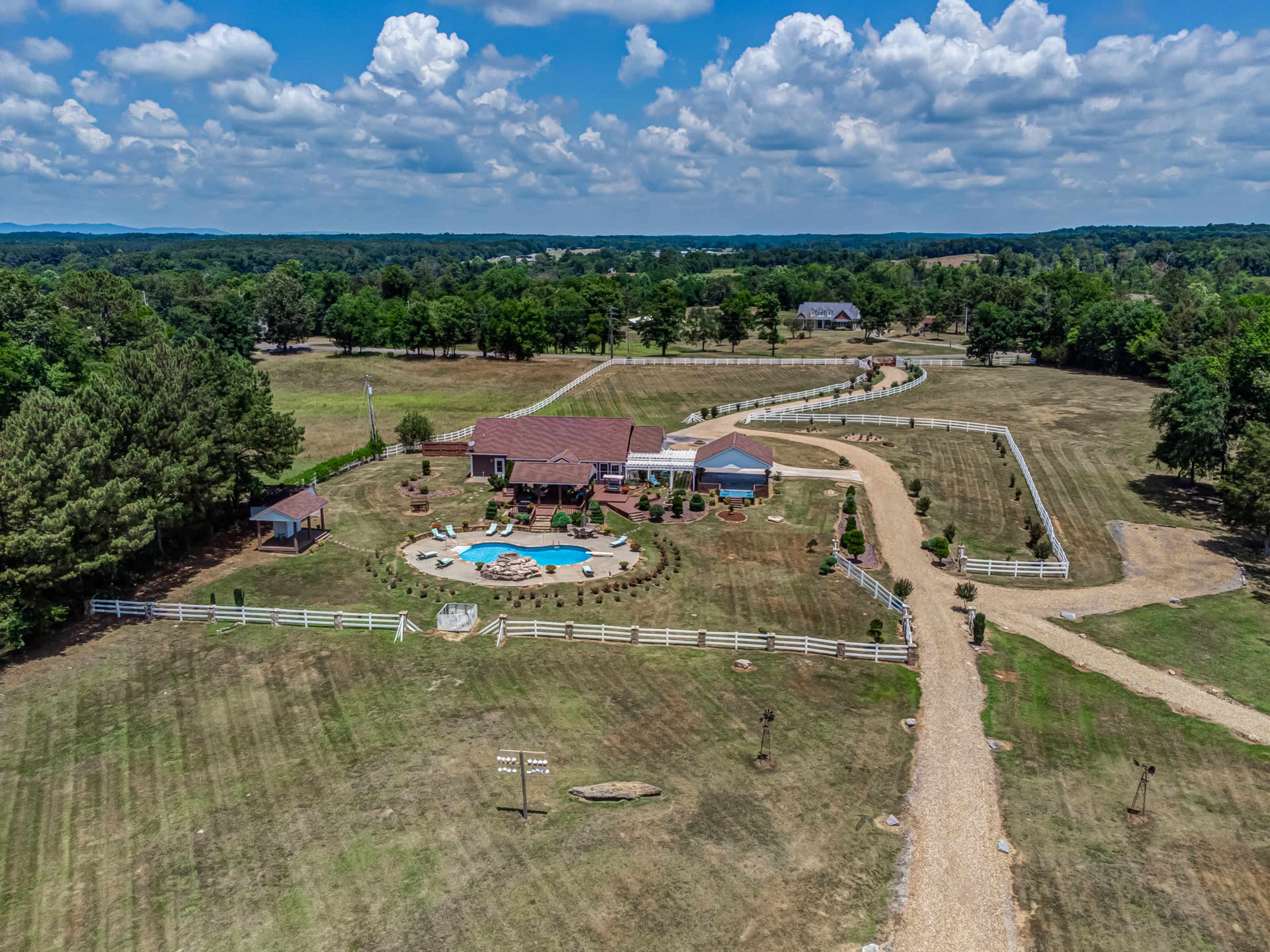 The image shows a large property with a house, a swimming pool, landscaped gardens, and a winding gravel driveway surrounded by open fields and trees.