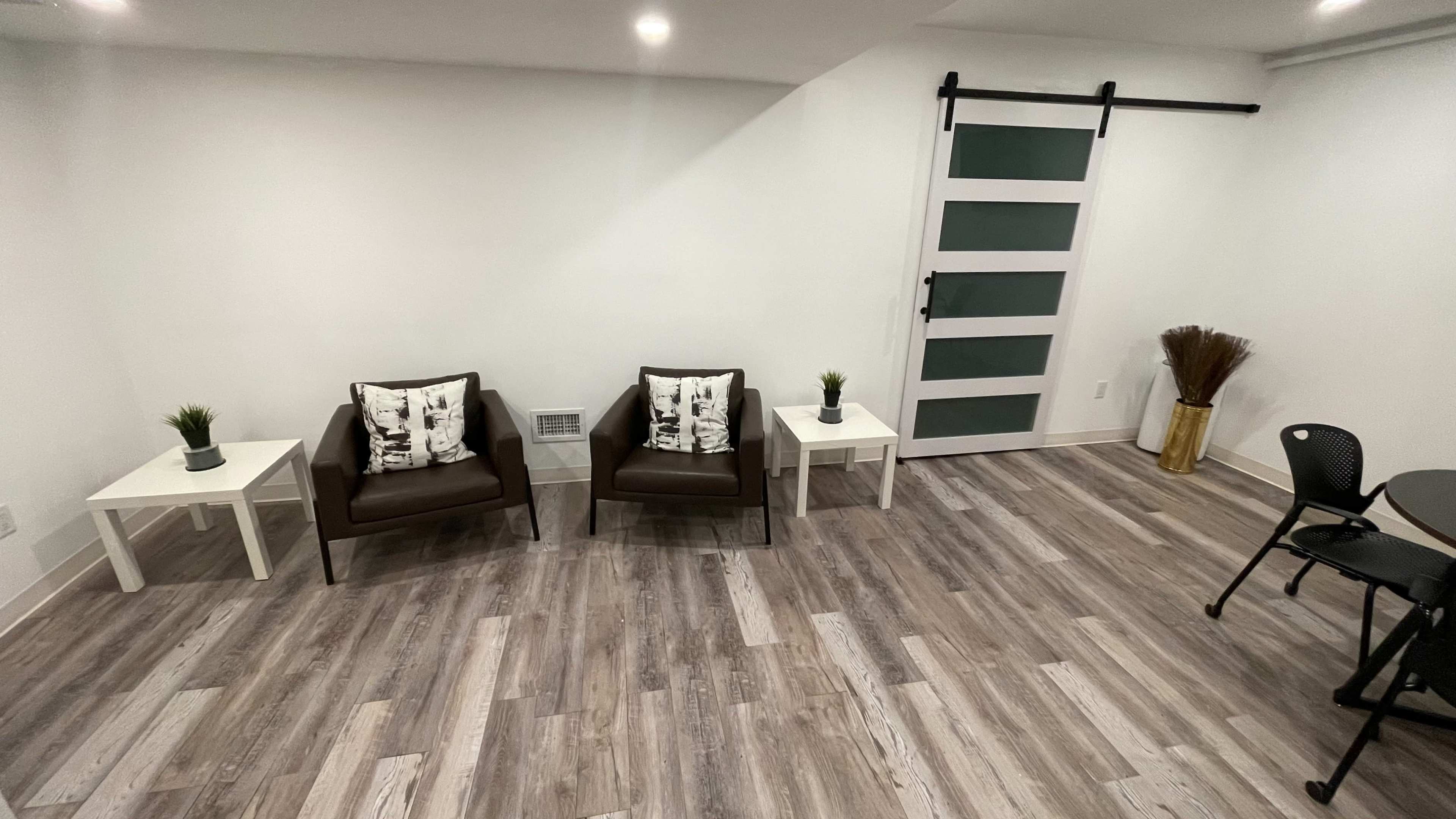 A minimalistic room with two brown chairs and small white tables, a black chair, and a patterned door in a well-lit space with wooden flooring.