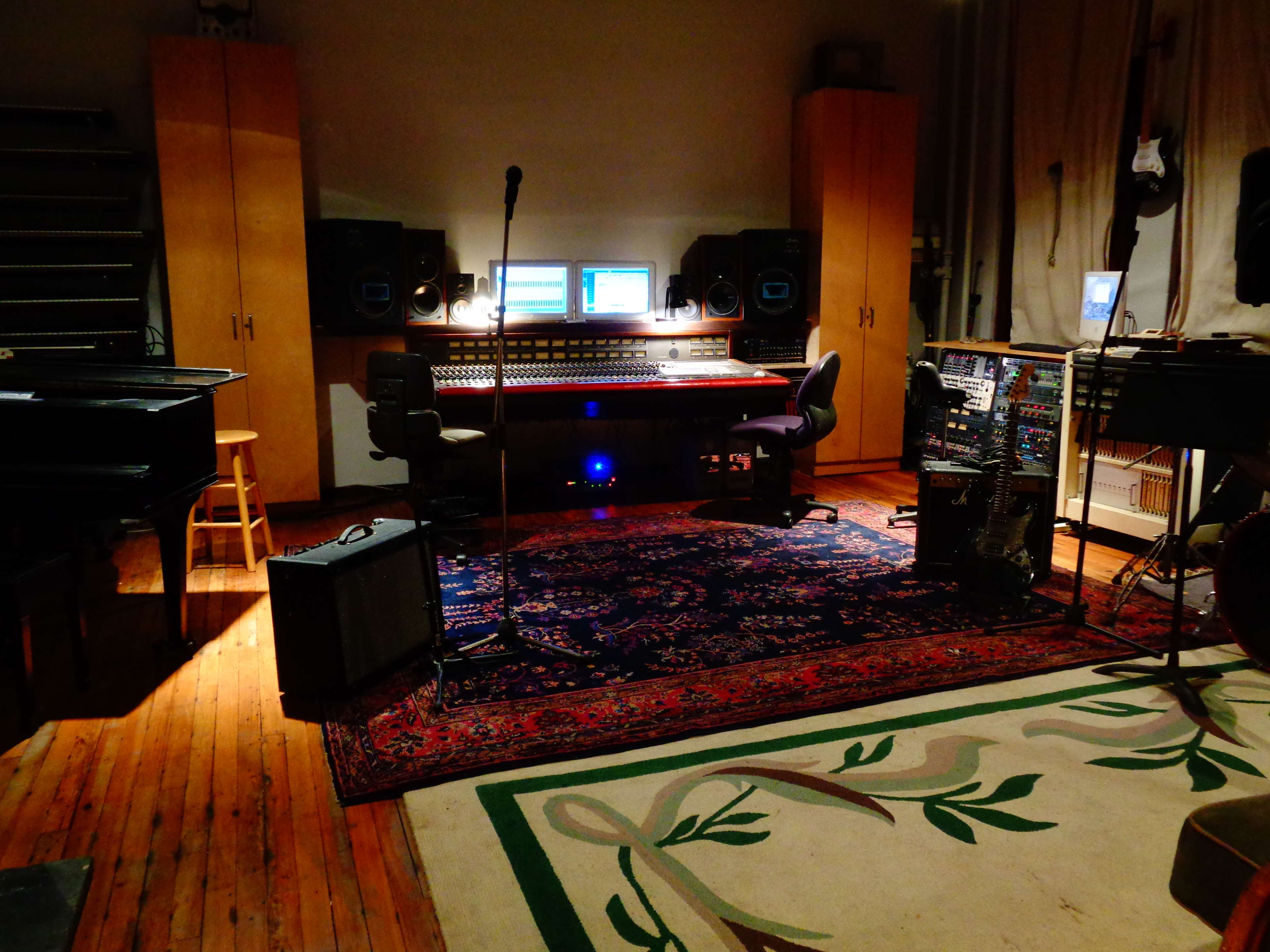 The image shows a music recording studio with various equipment, including a mixing console, monitors, microphones, and instruments, arranged around a patterned area rug.