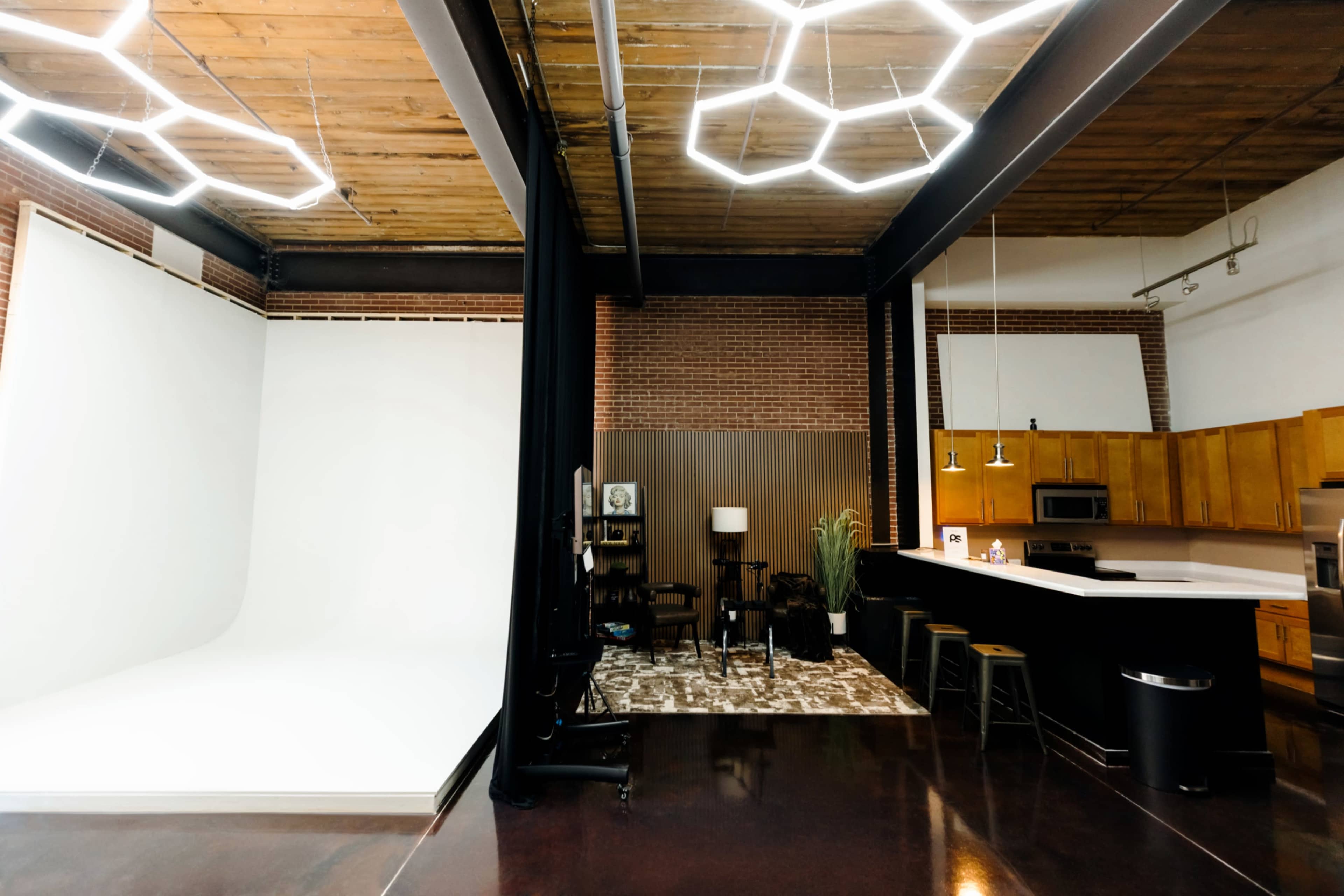 The image shows a spacious studio featuring a white backdrop on one side, a dark curtain, and a kitchen area with wooden cabinets and modern appliances on the other.