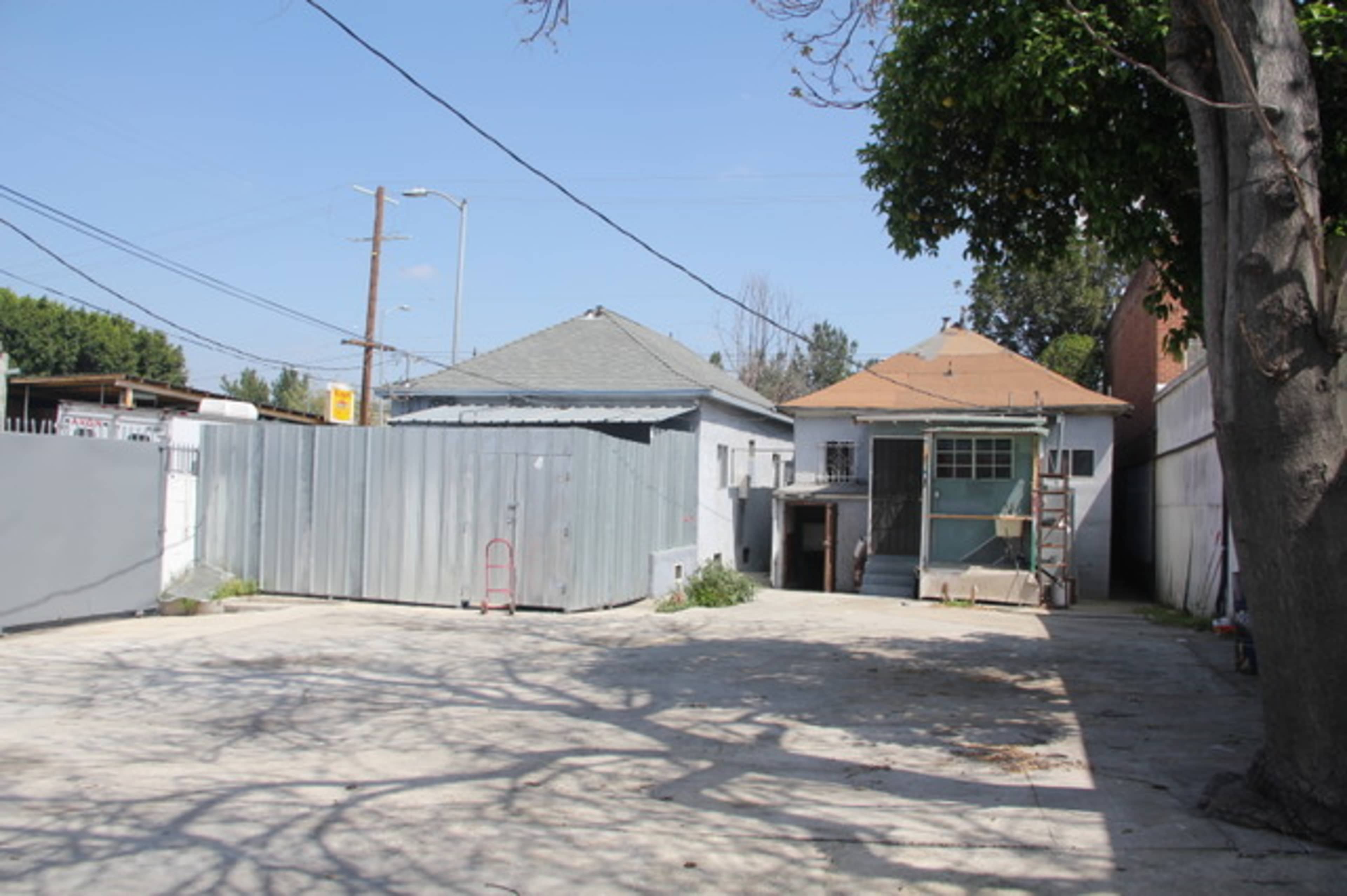 A vacant outdoor space with two houses in the background, surrounded by a fence and utility poles under a clear sky.