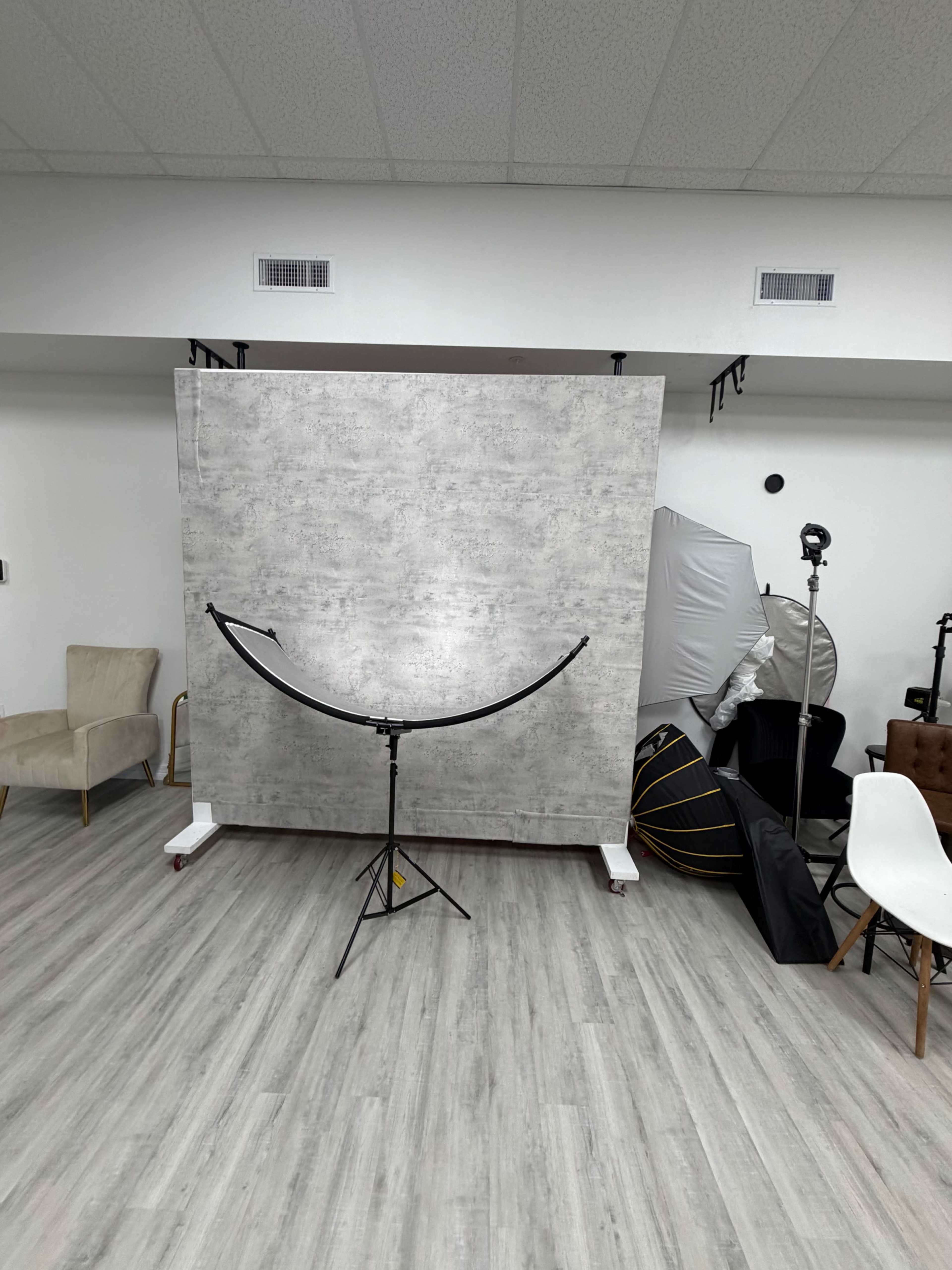The image depicts a photography studio setup featuring a textured backdrop, a curved light stand in front, and various studio equipment and furniture arranged in the space.