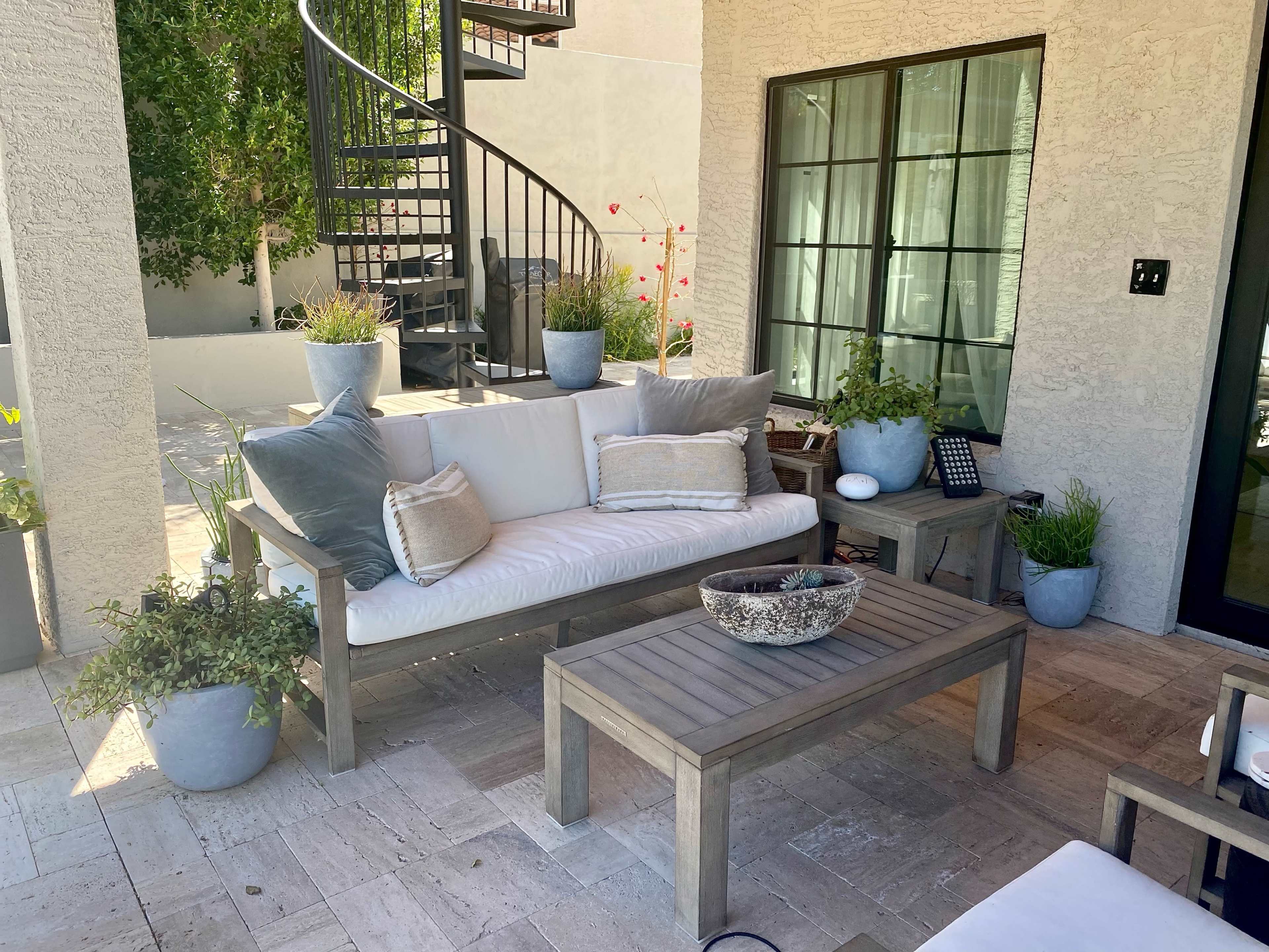 A patio area features a light-colored sofa with cushions, a wooden coffee table, and potted plants arranged around a stone floor.