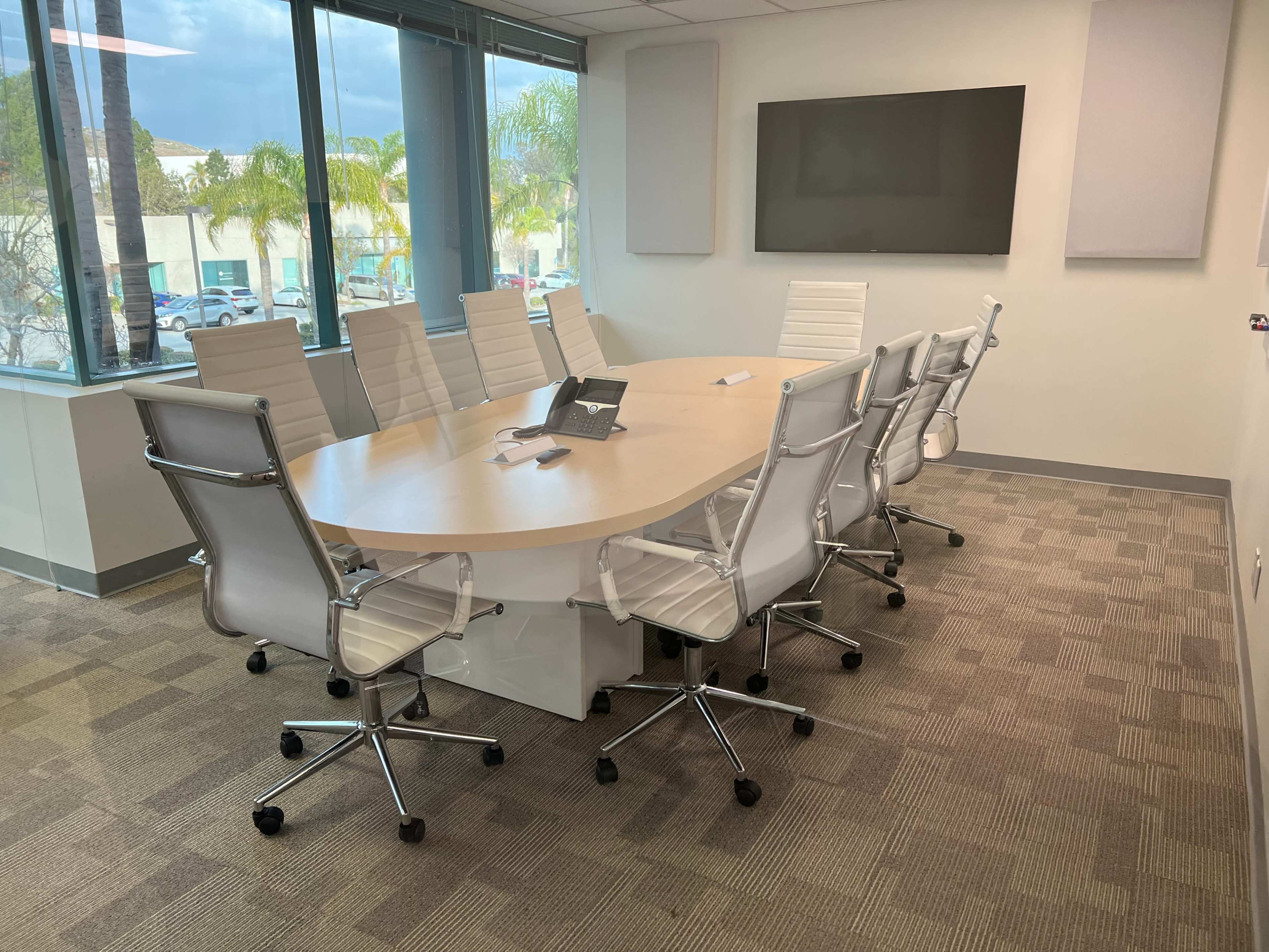 A modern conference room features a large oval table surrounded by white swivel chairs, with a large screen mounted on the wall and windows overlooking a view of palm trees.