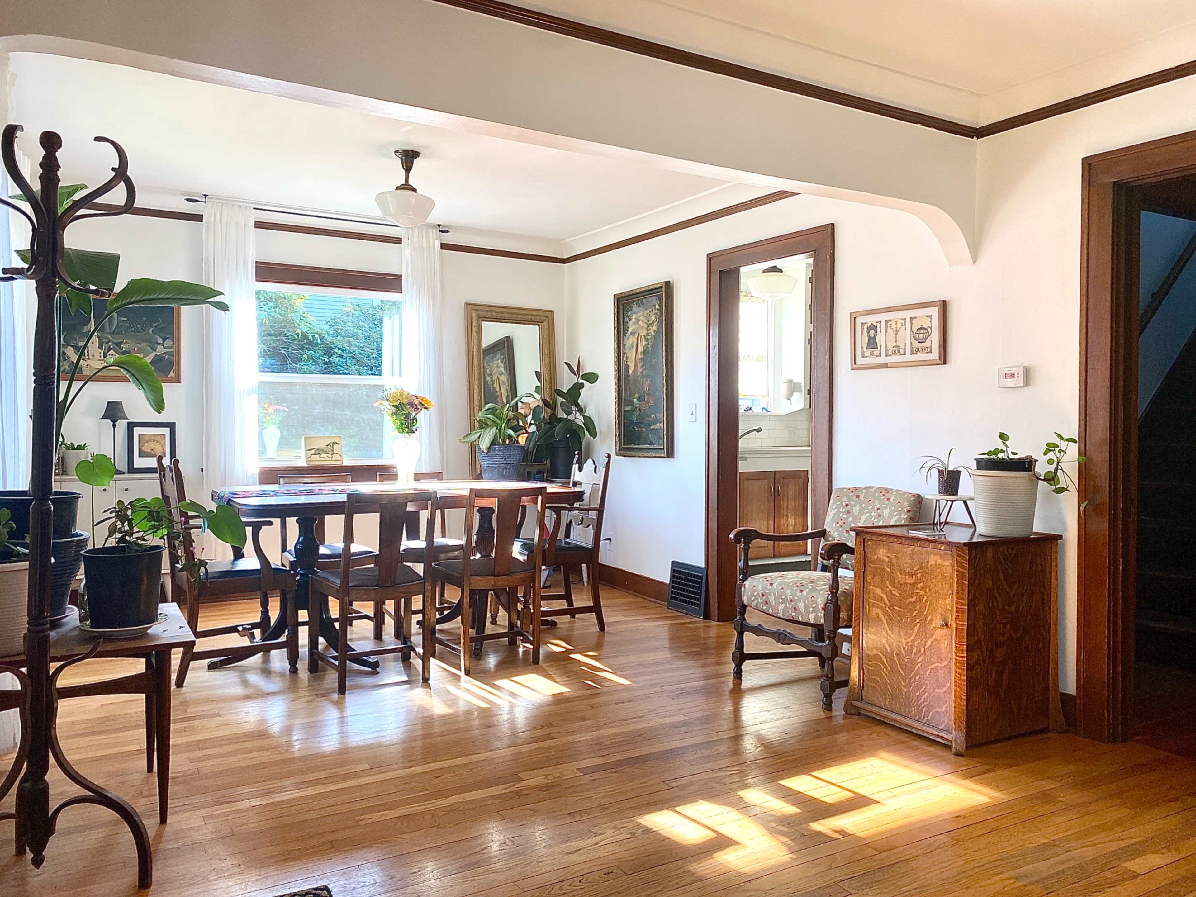 A bright dining area features a wooden table surrounded by chairs, with large windows and various houseplants enhancing the space.