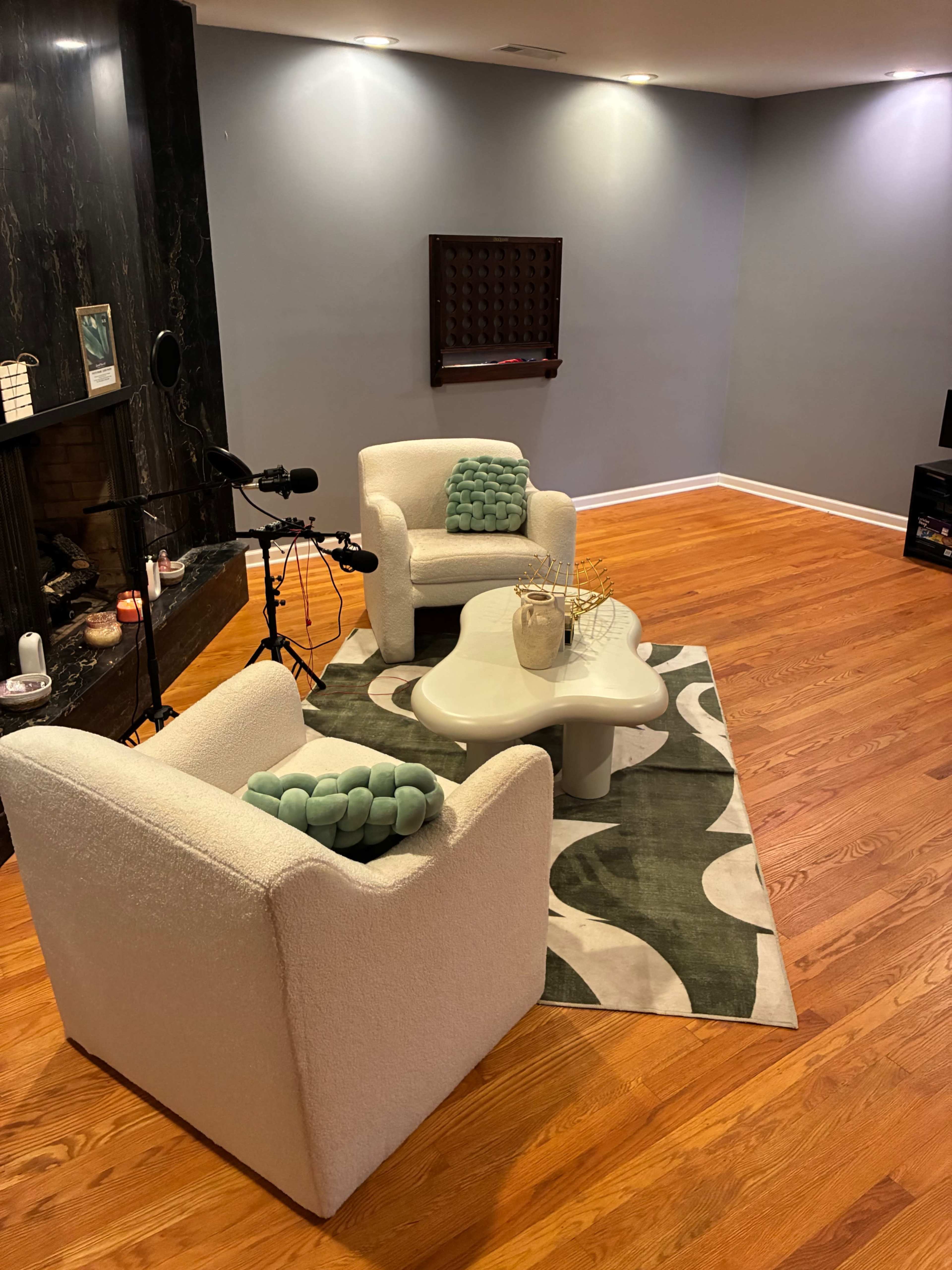 The scene features two white chairs with green pillows, a round coffee table, and a large patterned rug in a softly lit room with hardwood floors.