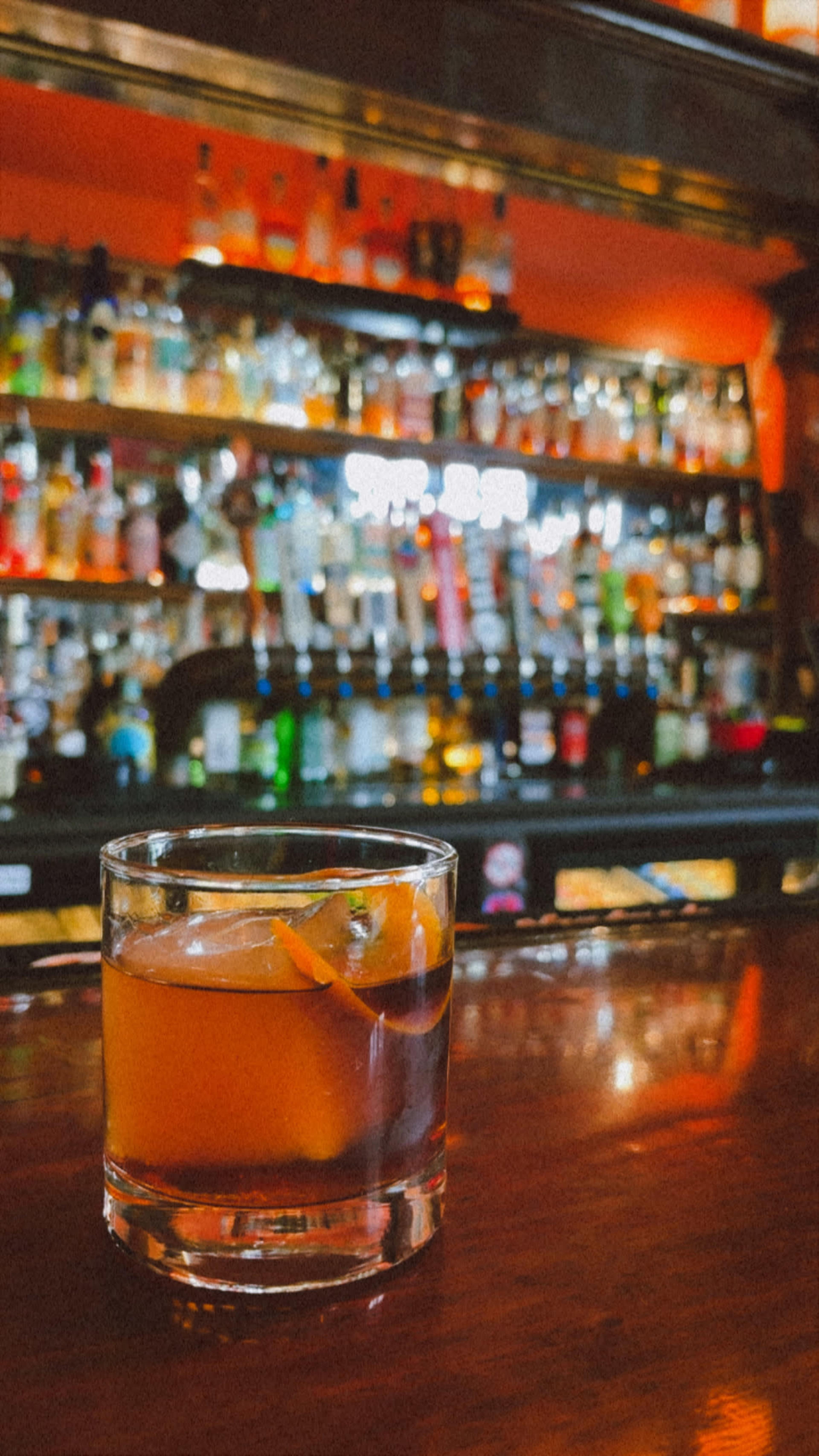 A glass of cocktail sits on a polished wooden bar top with a background of various bottles of liquor neatly arranged on shelves.