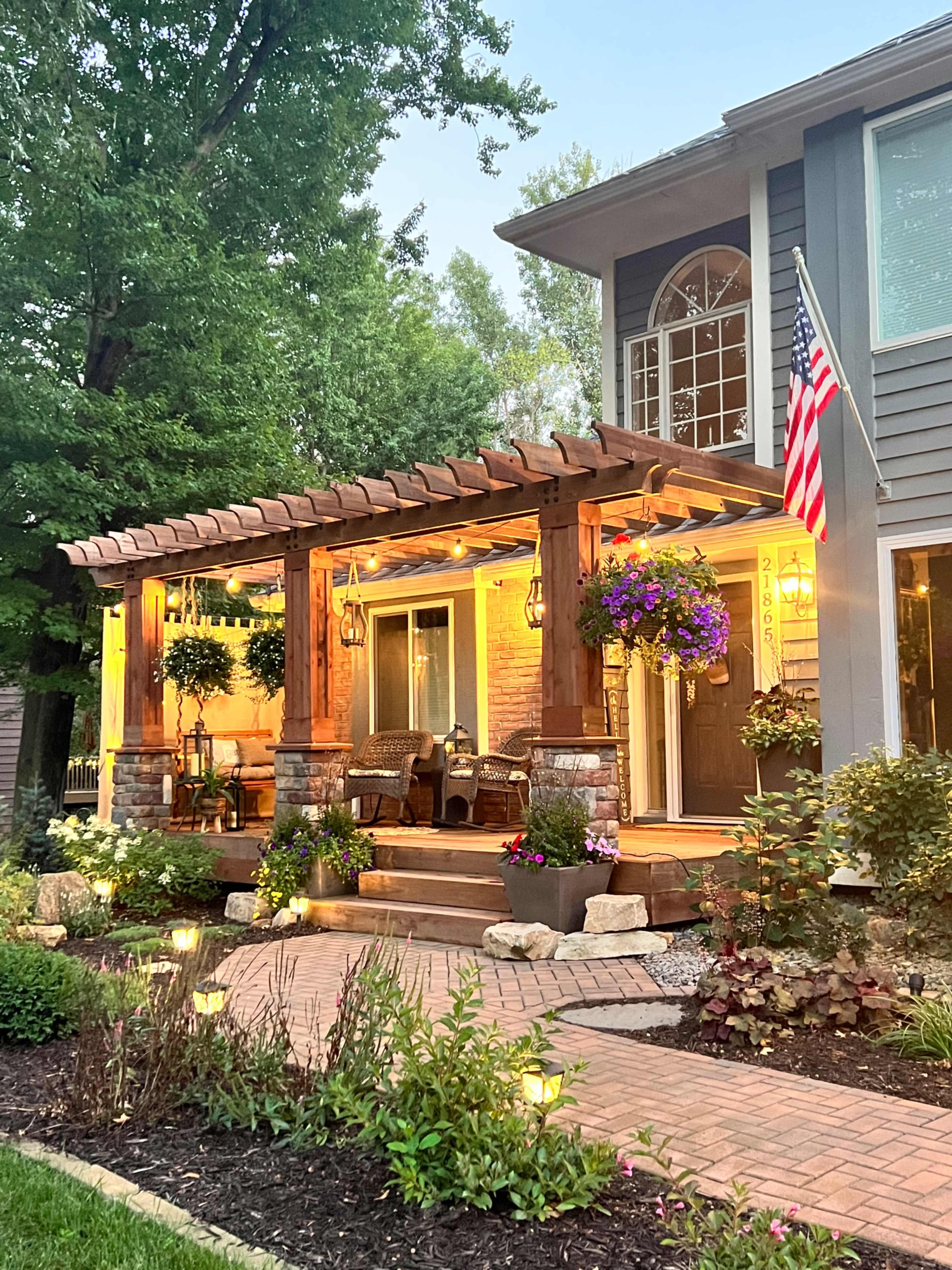 A wooden pergola with hanging baskets and two chairs leads to a well-lit entrance of a two-story home, adorned with American flags and surrounded by landscaped gardens.