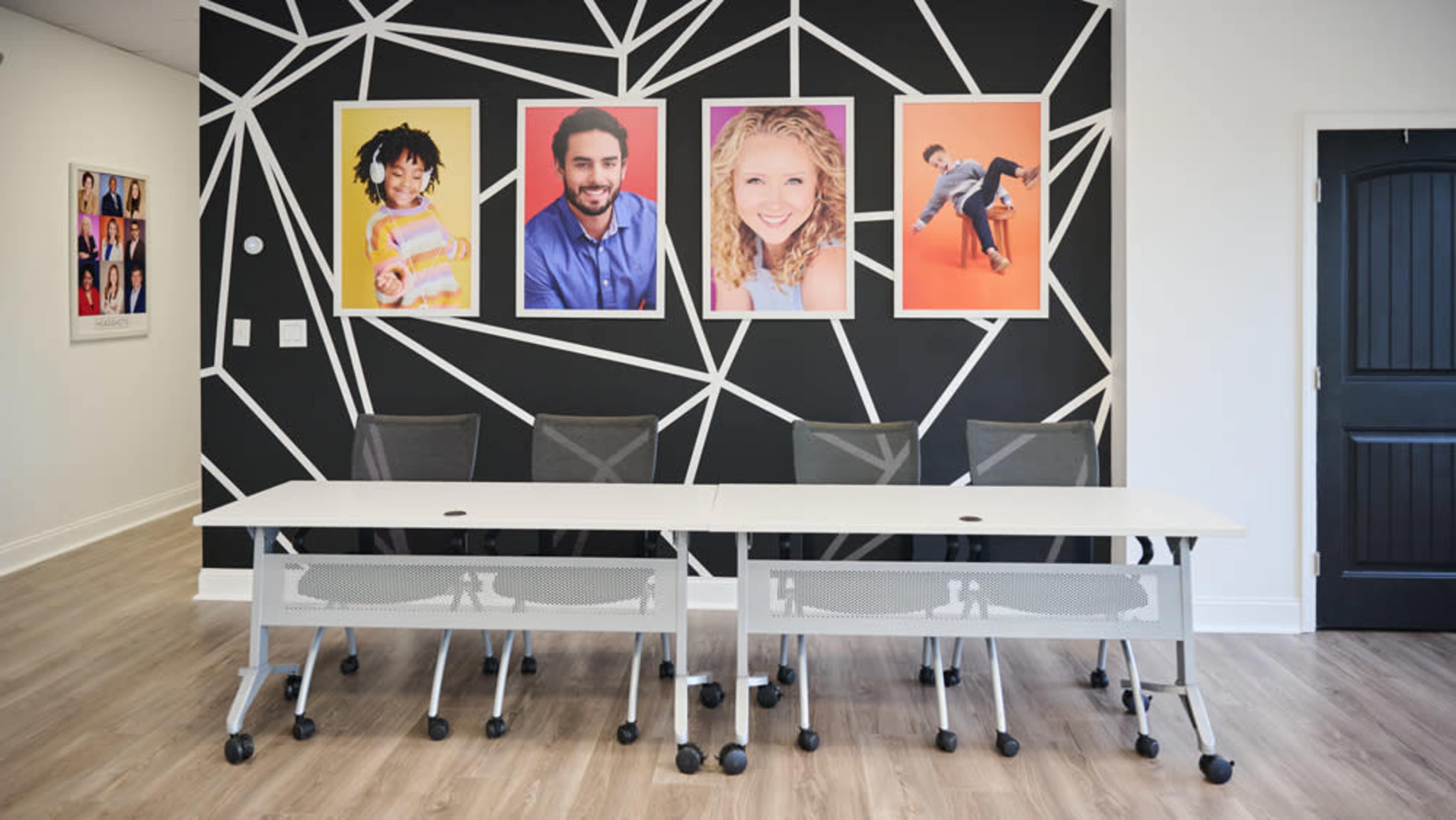 The image shows a modern conference room with a white table and rolling chairs, featuring a geometric black wall mural adorned with colorful portraits.