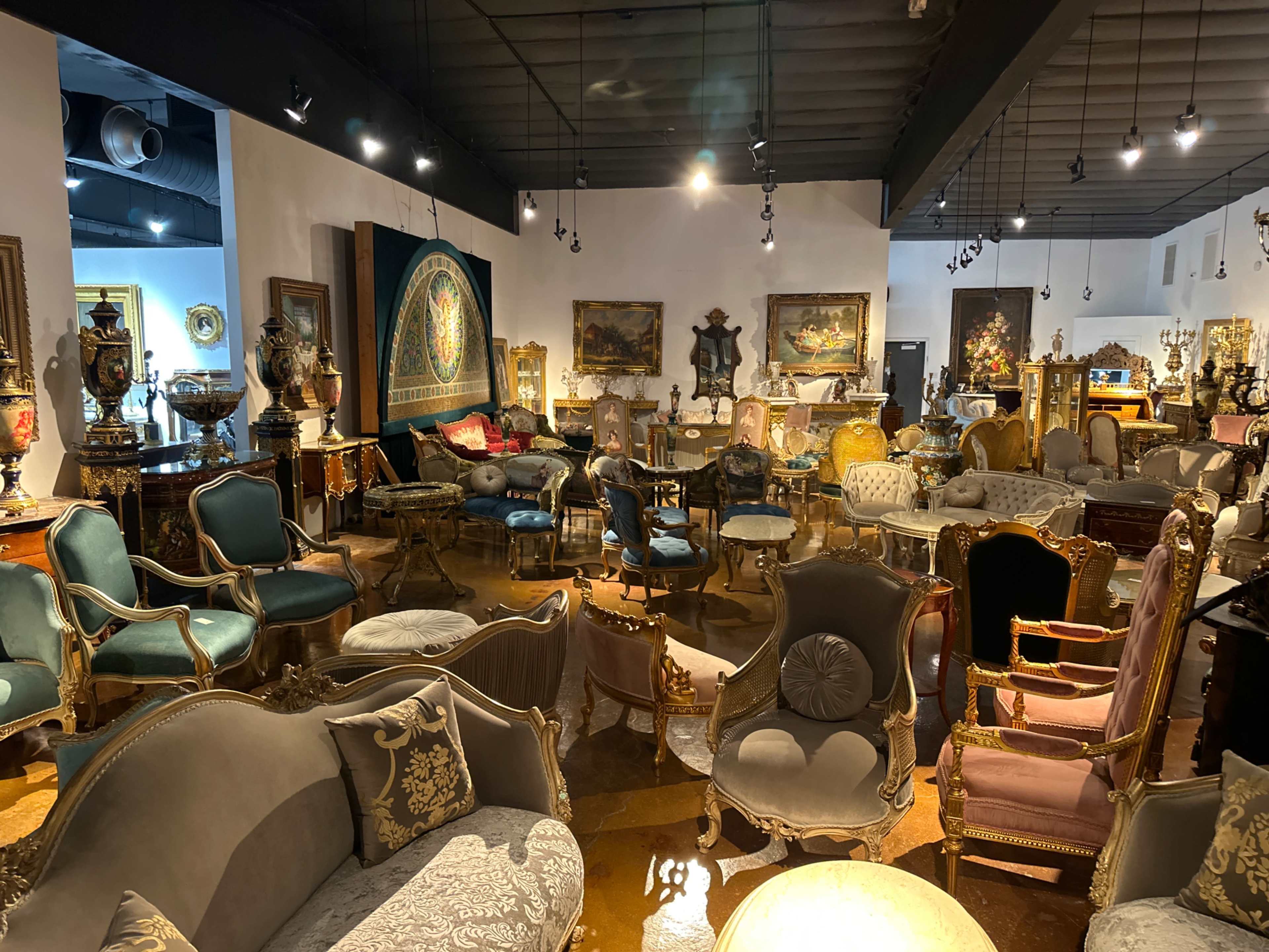 A spacious antique furniture showroom filled with various ornate chairs, sofas, and decorative artworks arranged throughout the room.