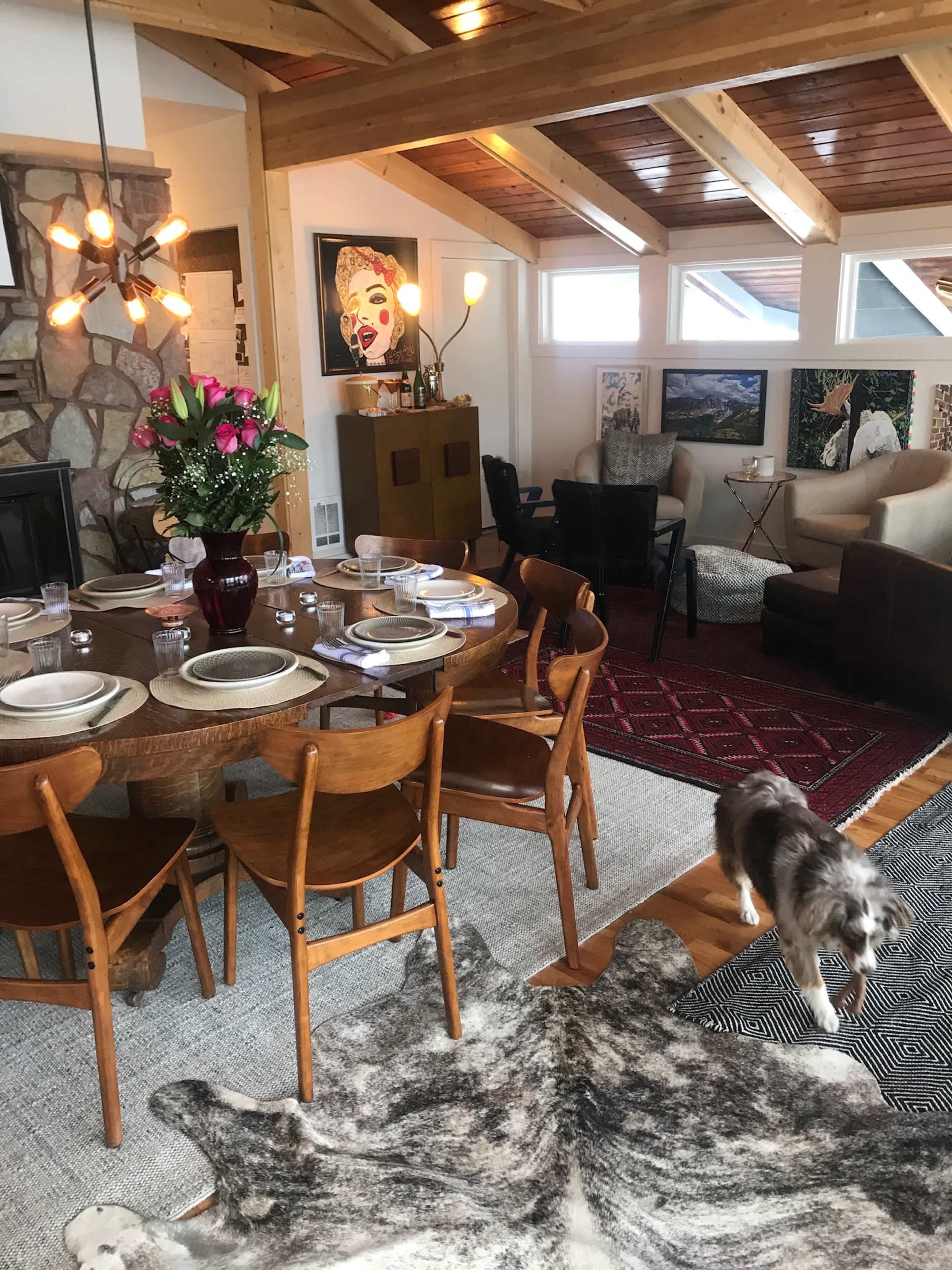 A spacious living and dining area features a round wooden table set for a meal, surrounded by chairs, with a dog walking on a patterned rug.