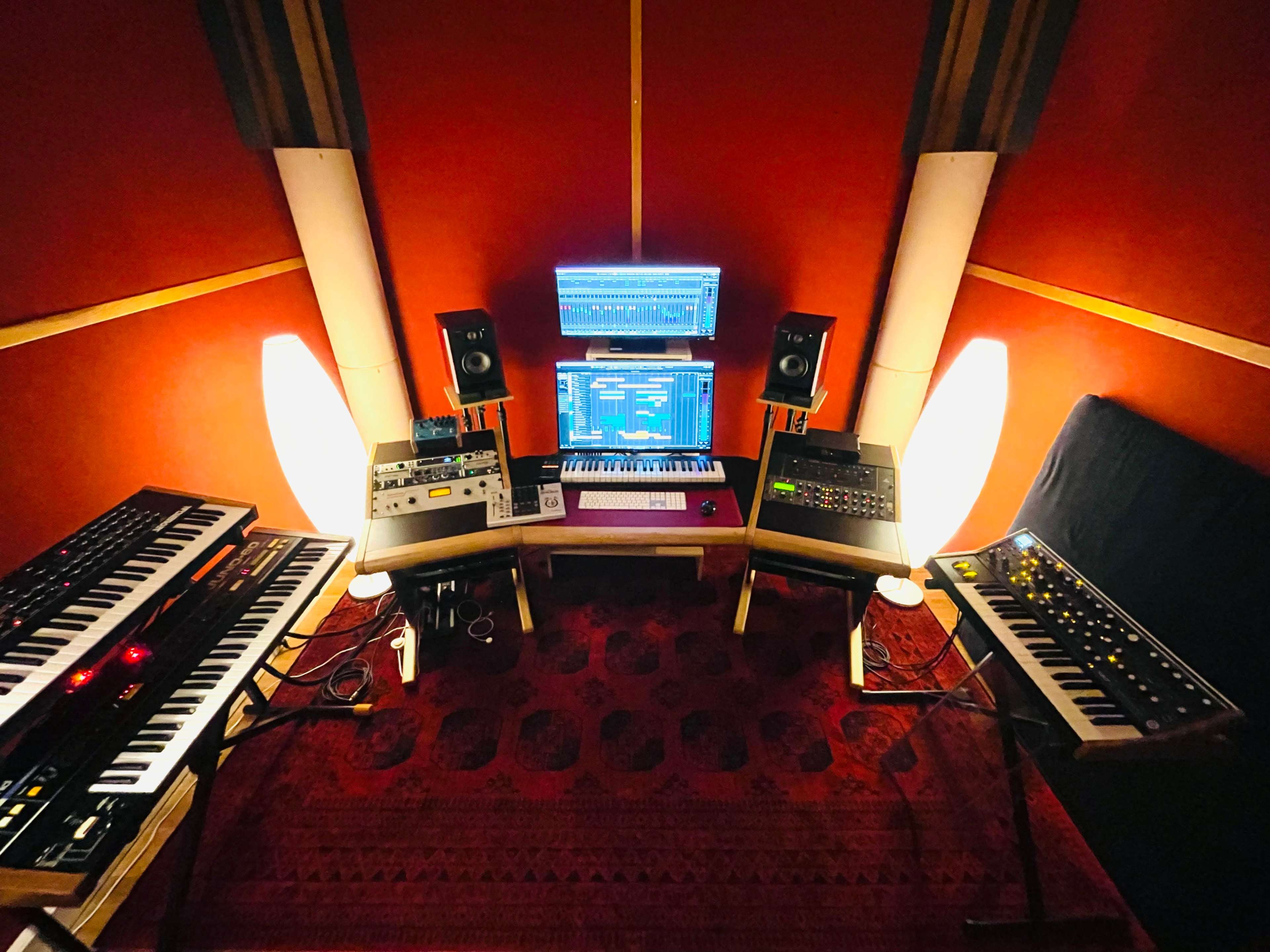 A music production setup featuring multiple synthesizers, a computer screen, and speakers arranged in a small studio space with a red backdrop and a patterned rug.