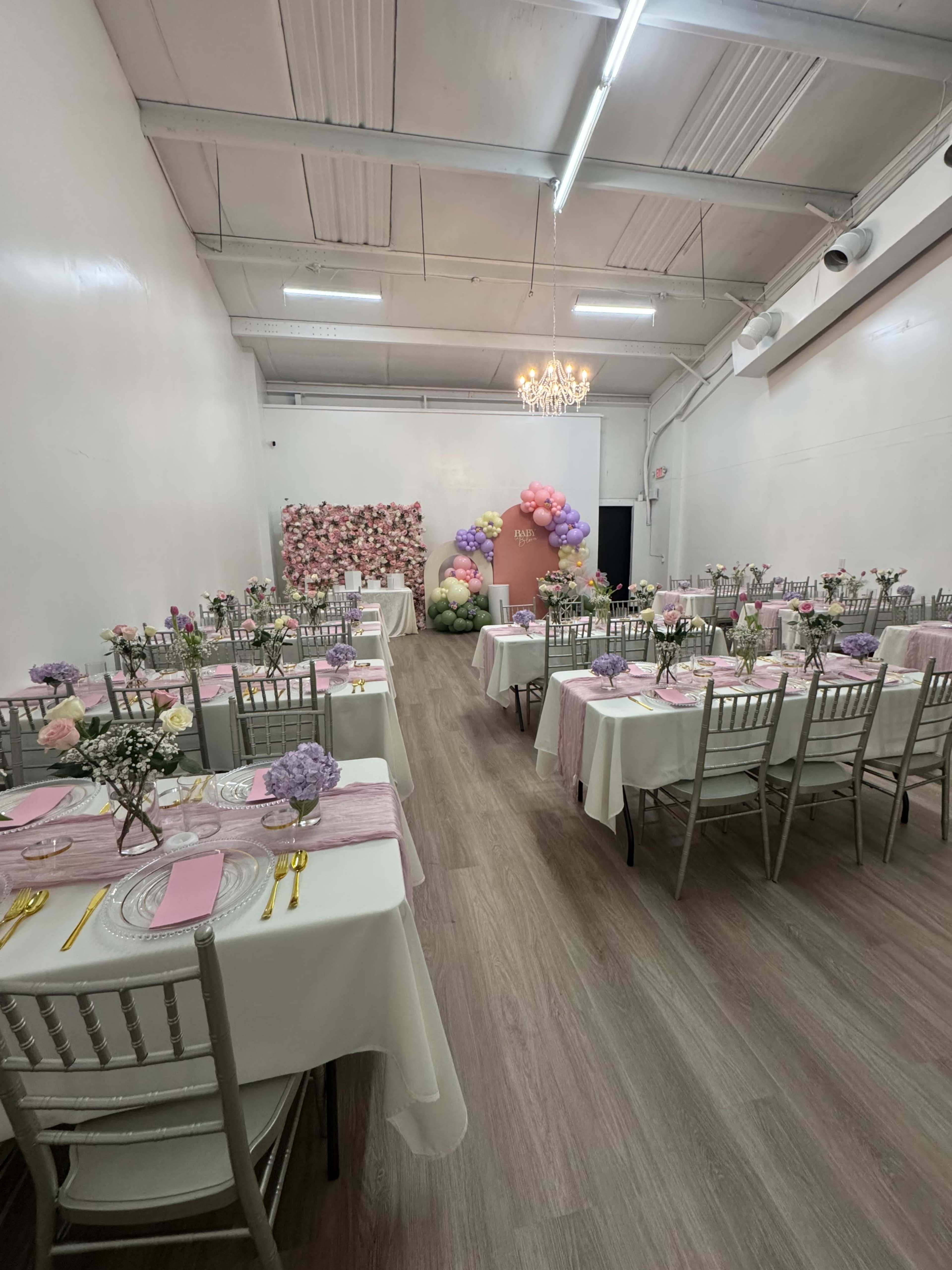 The image shows a decorated event space with tables set for a celebration, featuring floral arrangements and a colorful balloon backdrop.