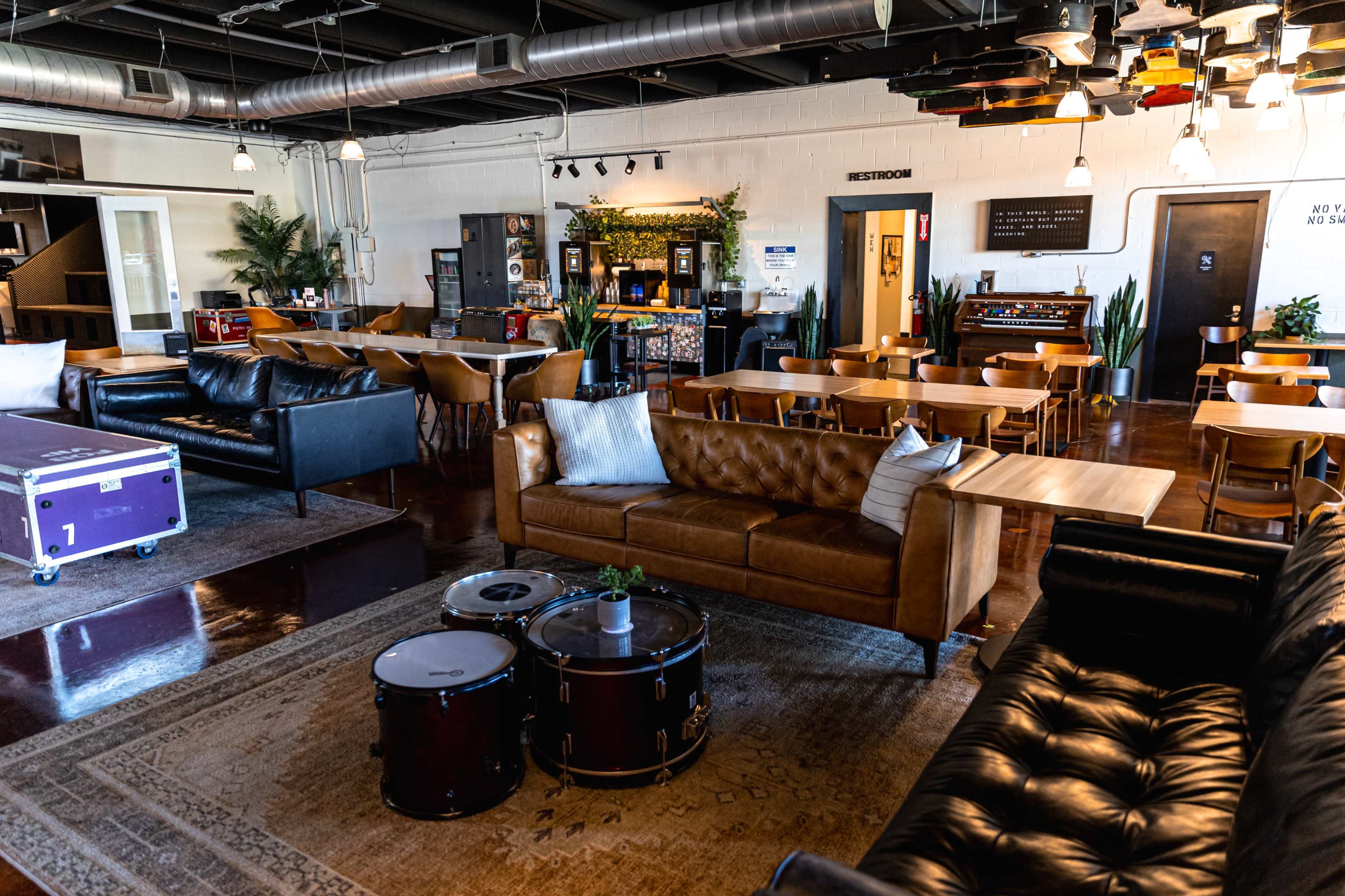 The image shows a spacious lounge area featuring leather sofas, wooden tables, and a drum set on a rug, with a dining area visible in the background.