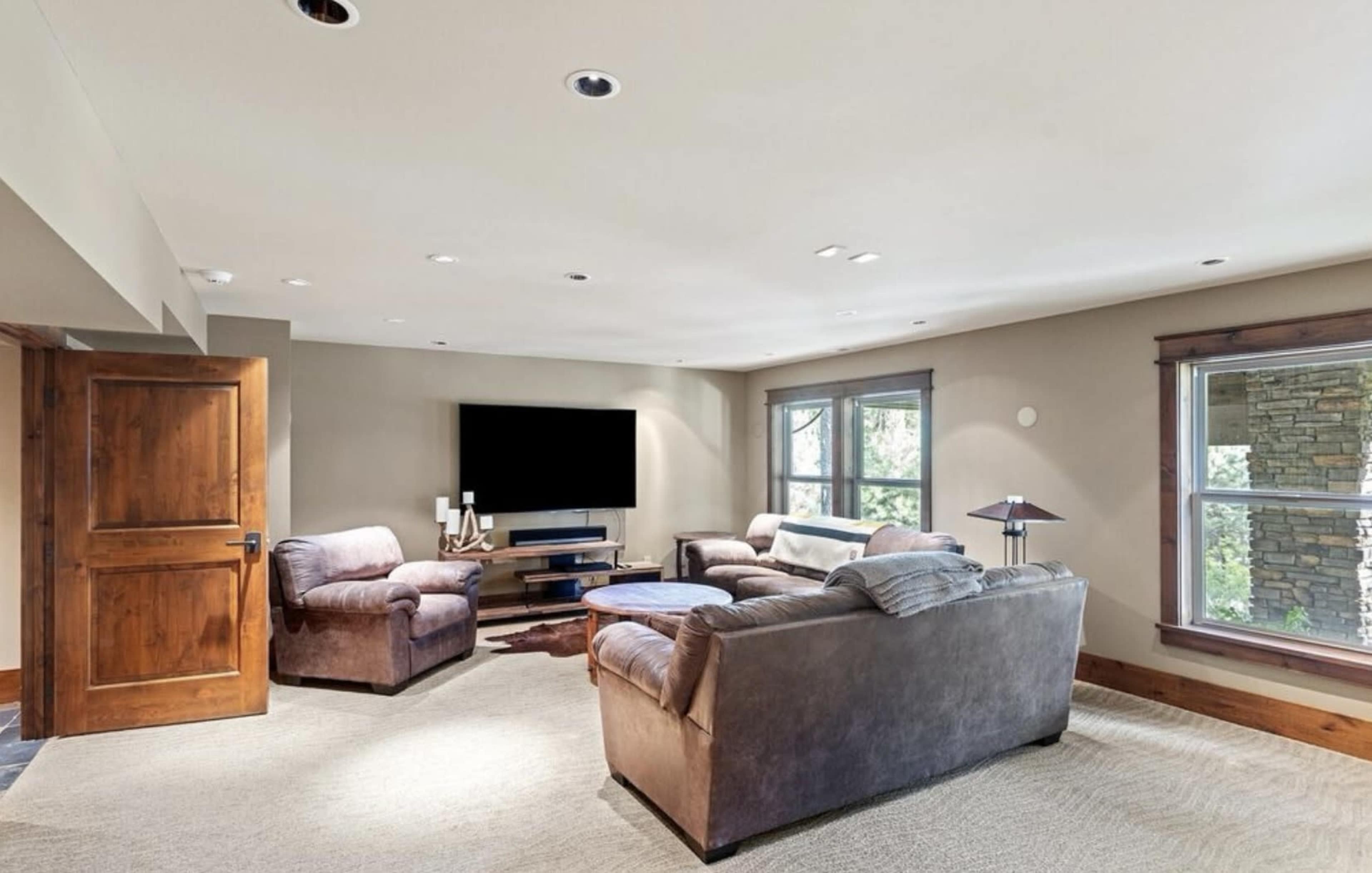 The image shows a cozy living room with two brown sofas, a television mounted on the wall, and large windows letting in natural light.