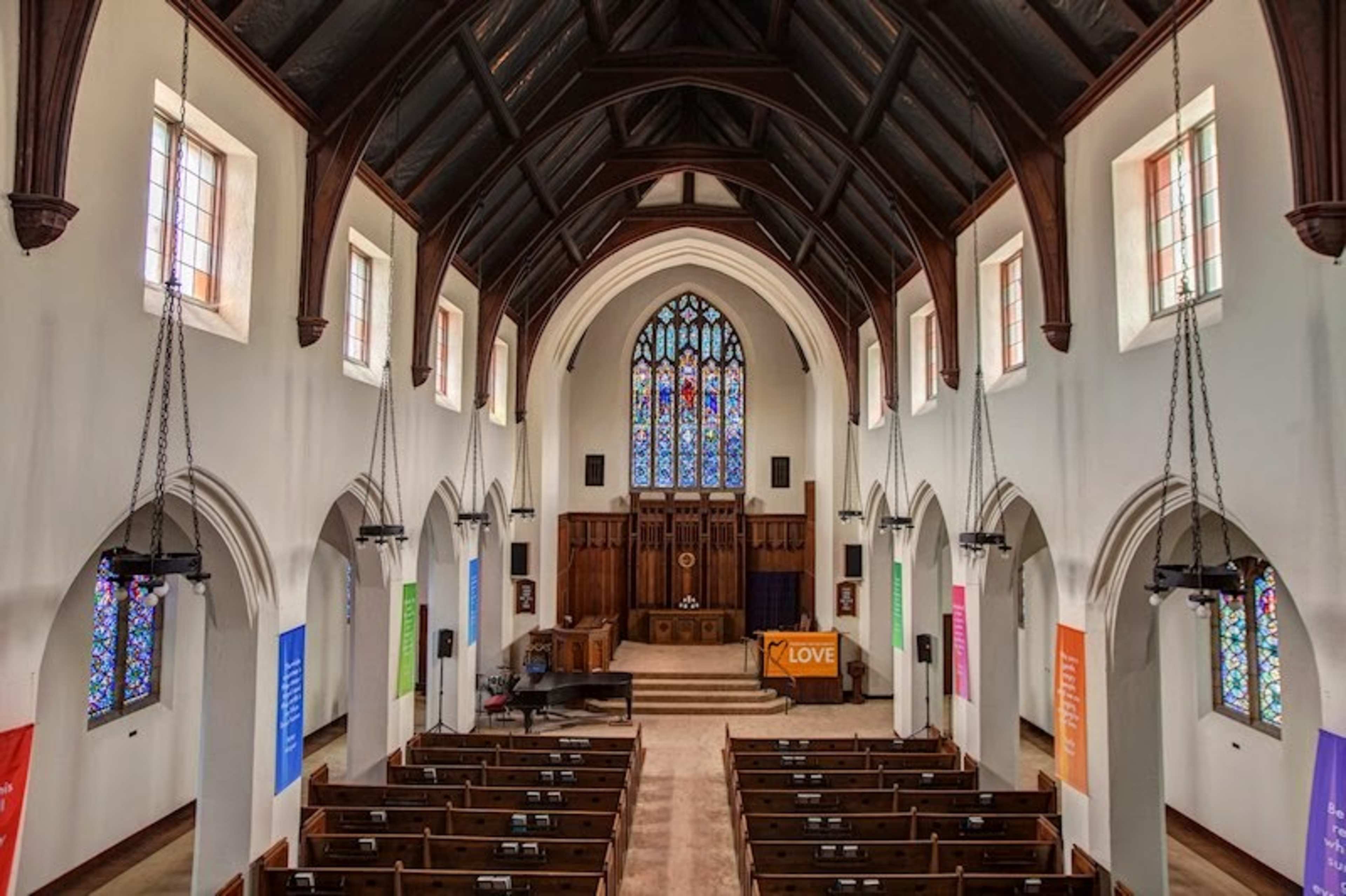 The interior of a church features a high vaulted ceiling, stained glass windows, and wooden benches arranged in rows facing a stage.