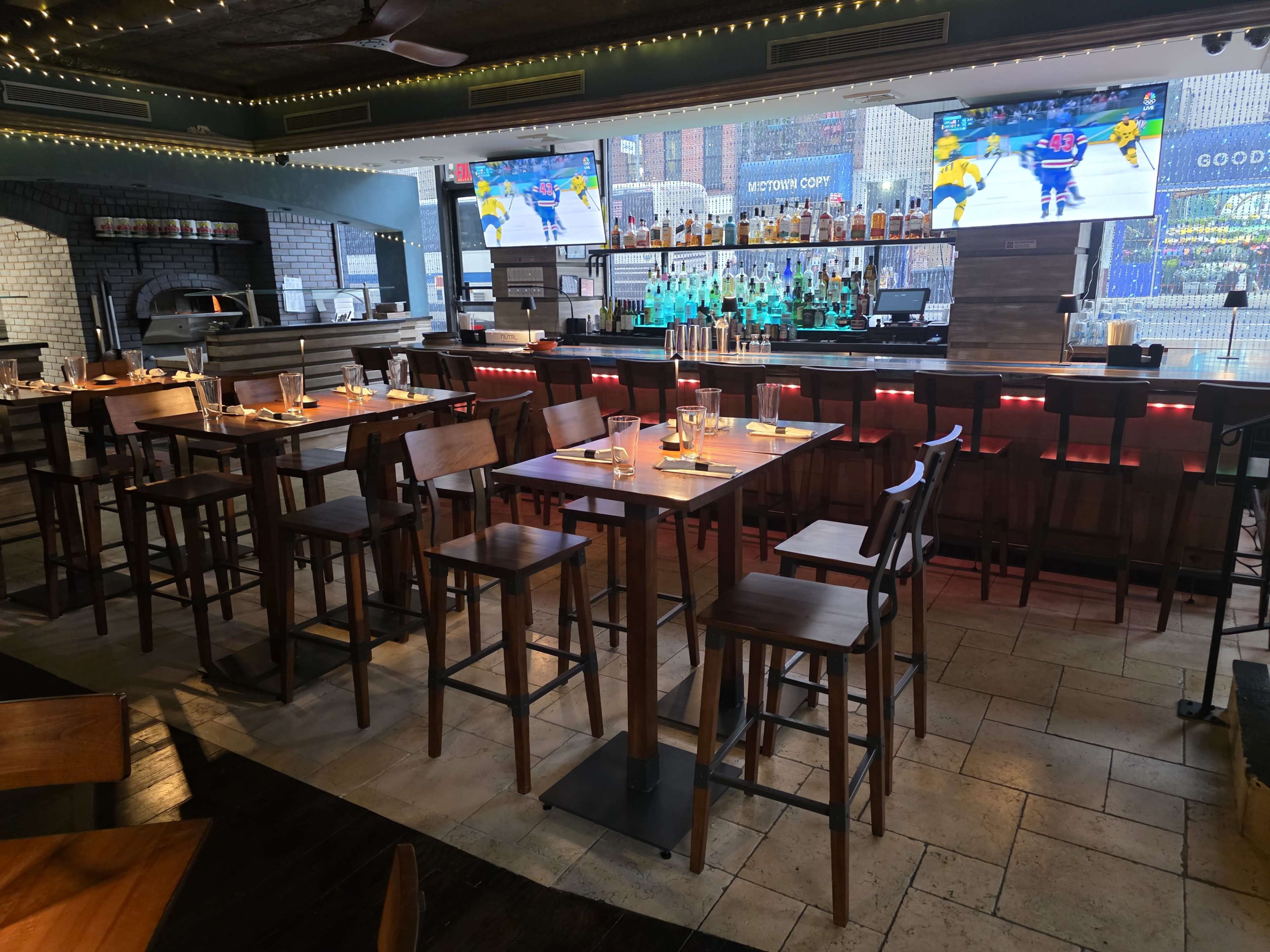 The image shows a modern bar area with high wooden stools, a well-stocked bar featuring multiple bottles, and large screens displaying a sports game in a spacious establishment.