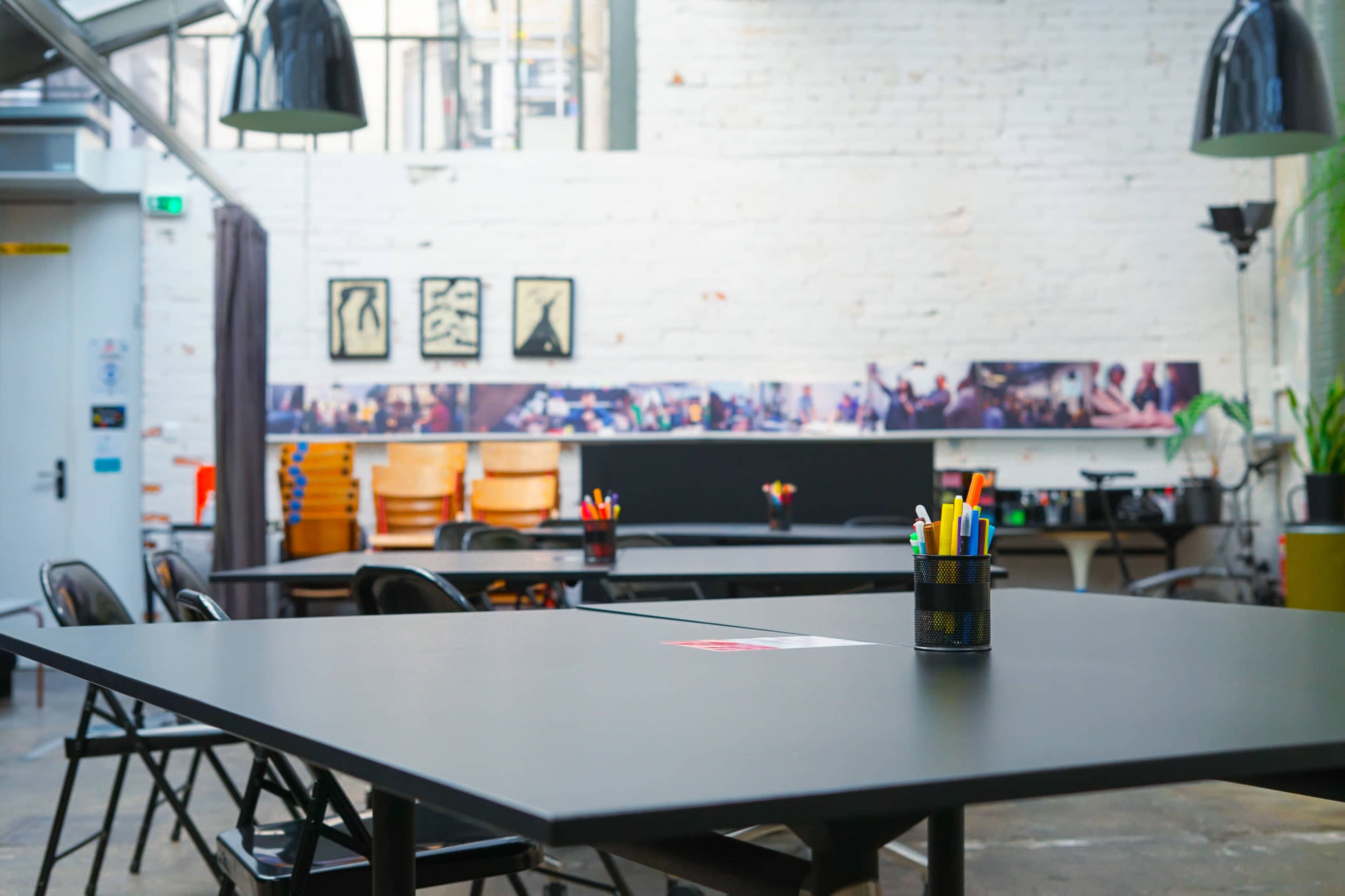A bright, open workspace features black tables, chairs, and a collection of colorful pens in a container on one of the tables.