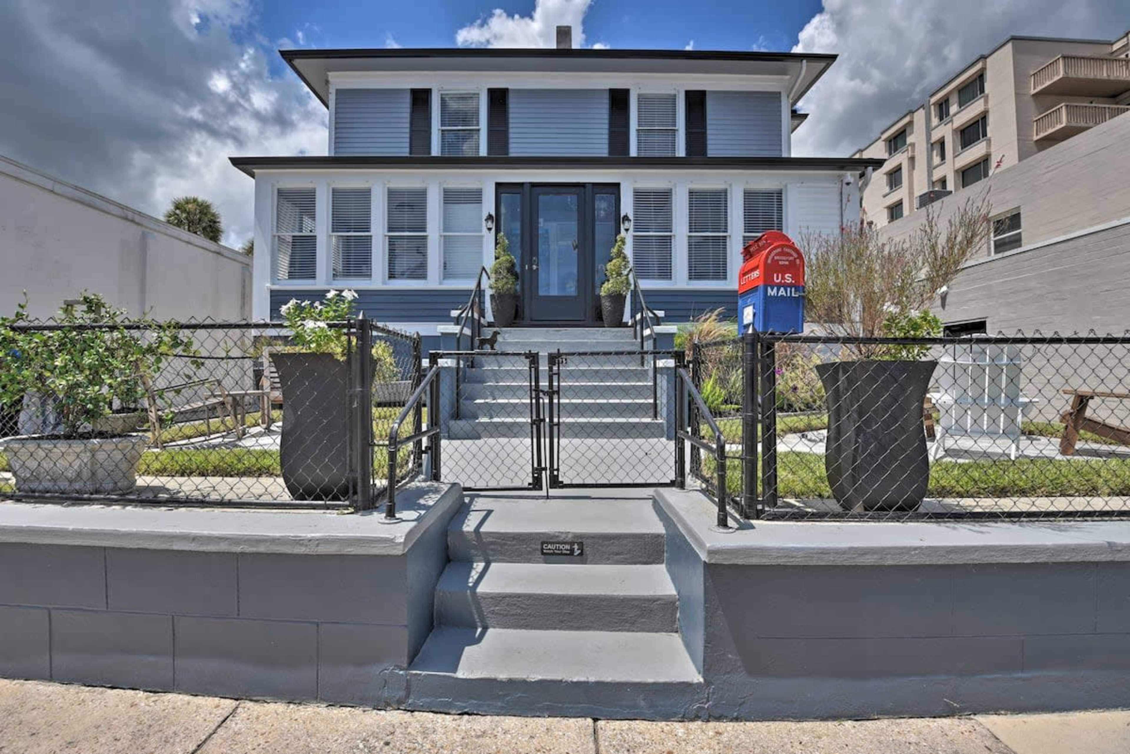 A two-story blue house with white trim features a set of steps leading to a central front door, flanked by large planters and a red mailbox at the entrance.