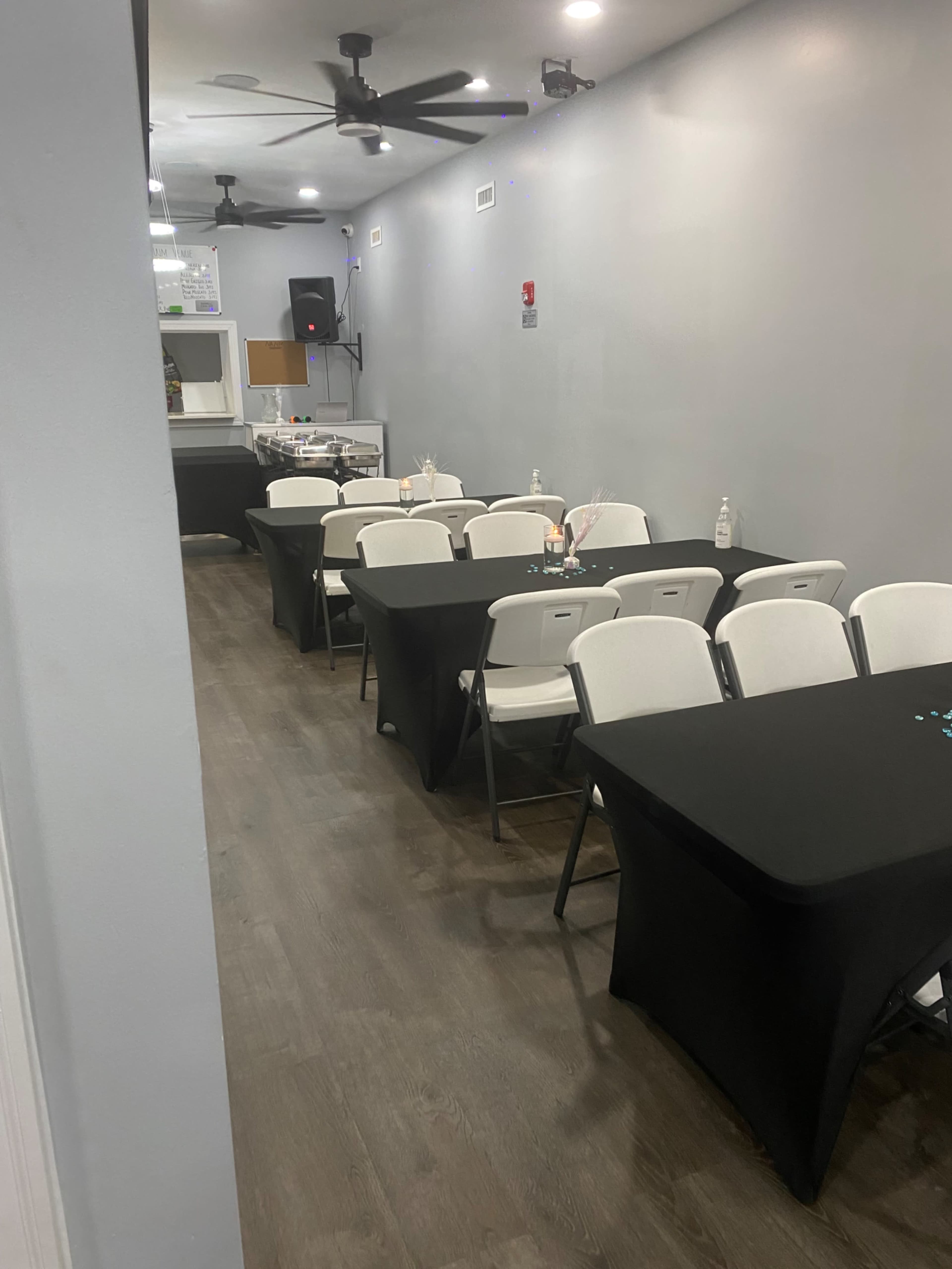 The image shows a narrow room with several rows of tables covered in black tablecloths and white folding chairs lined up, with a speaker system and speakers at one end.