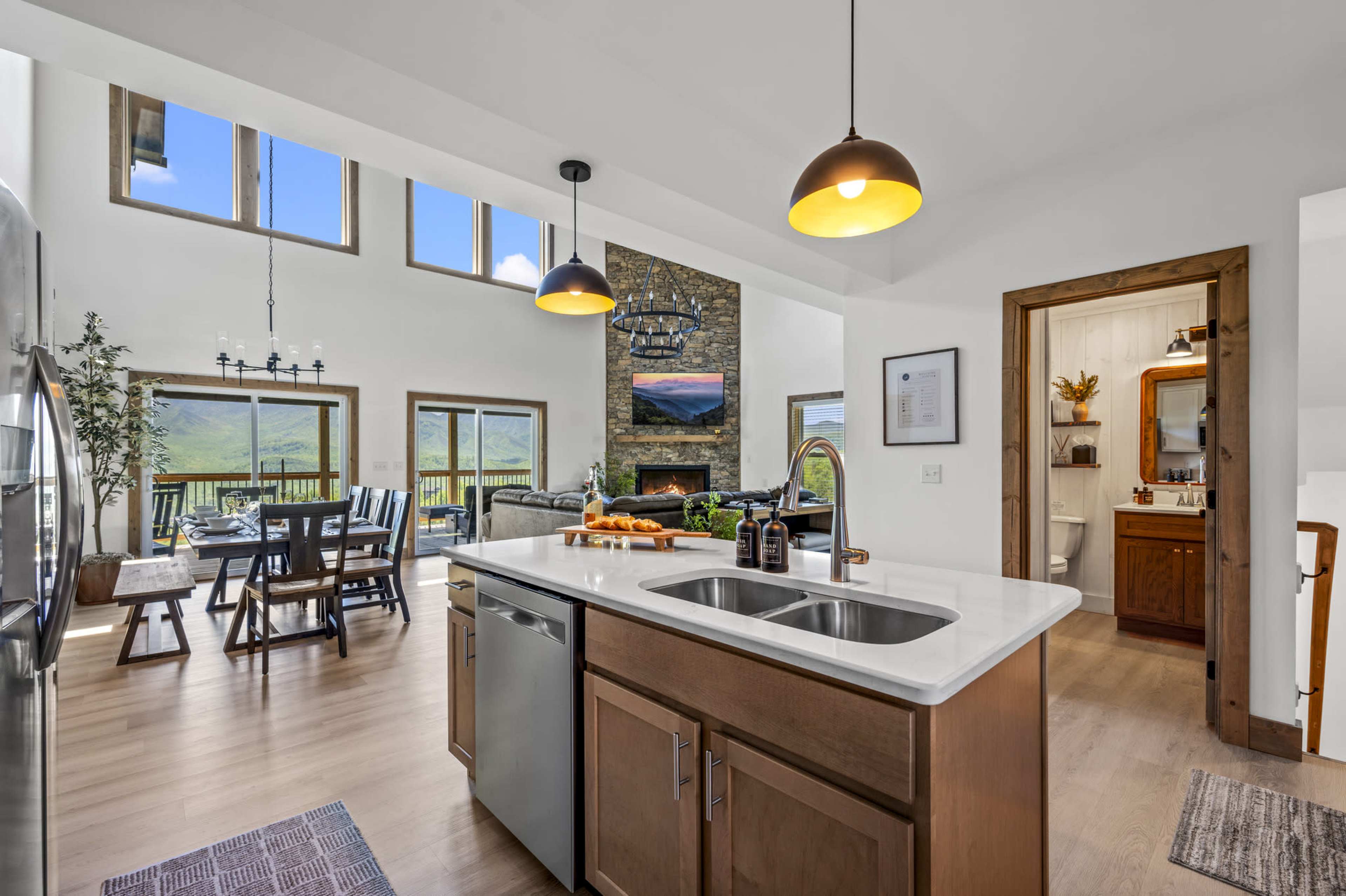 The image shows a modern kitchen with a central island, open dining area, and large windows providing views of the outdoors.