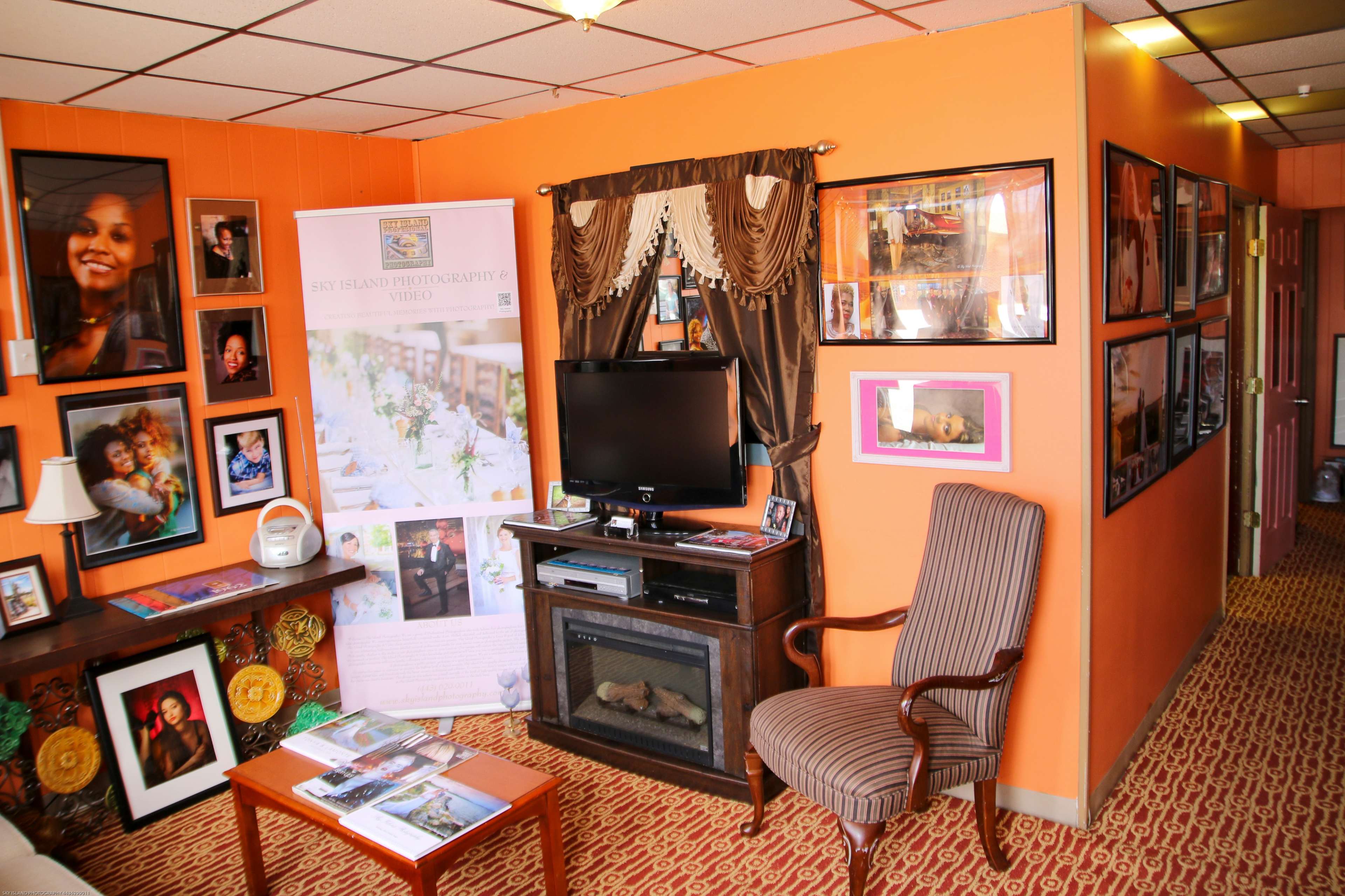 A cozy interior room with orange walls, featuring a television, a decorative chair, framed photographs, and a banner for a photography business.