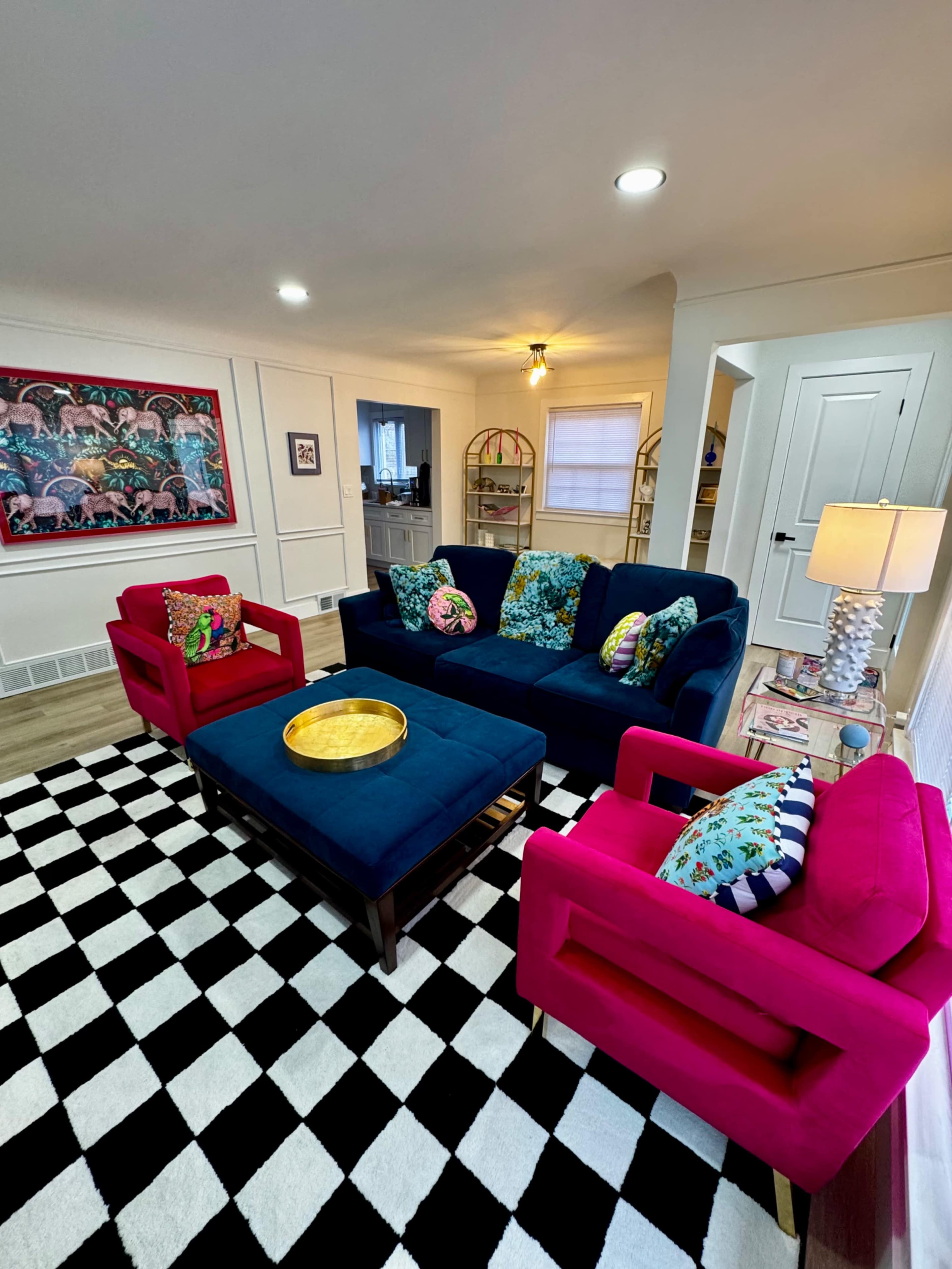 A living room features a navy blue sofa and two bright pink armchairs, with a black and white checkered rug and decorative pillows on the furniture.