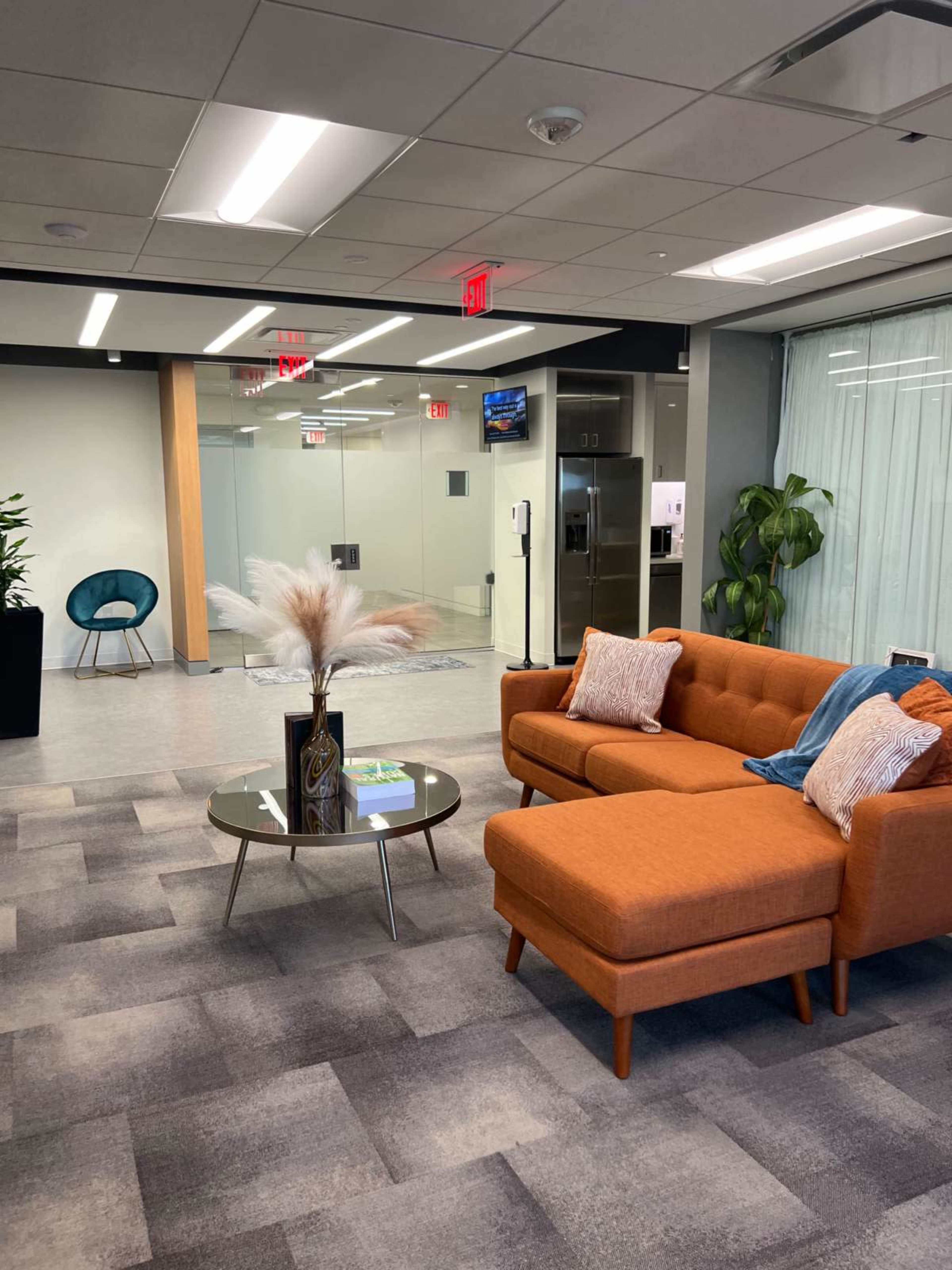 A modern office space features an orange sectional sofa, a glass coffee table with decorative items, and large windows with sheer curtains.