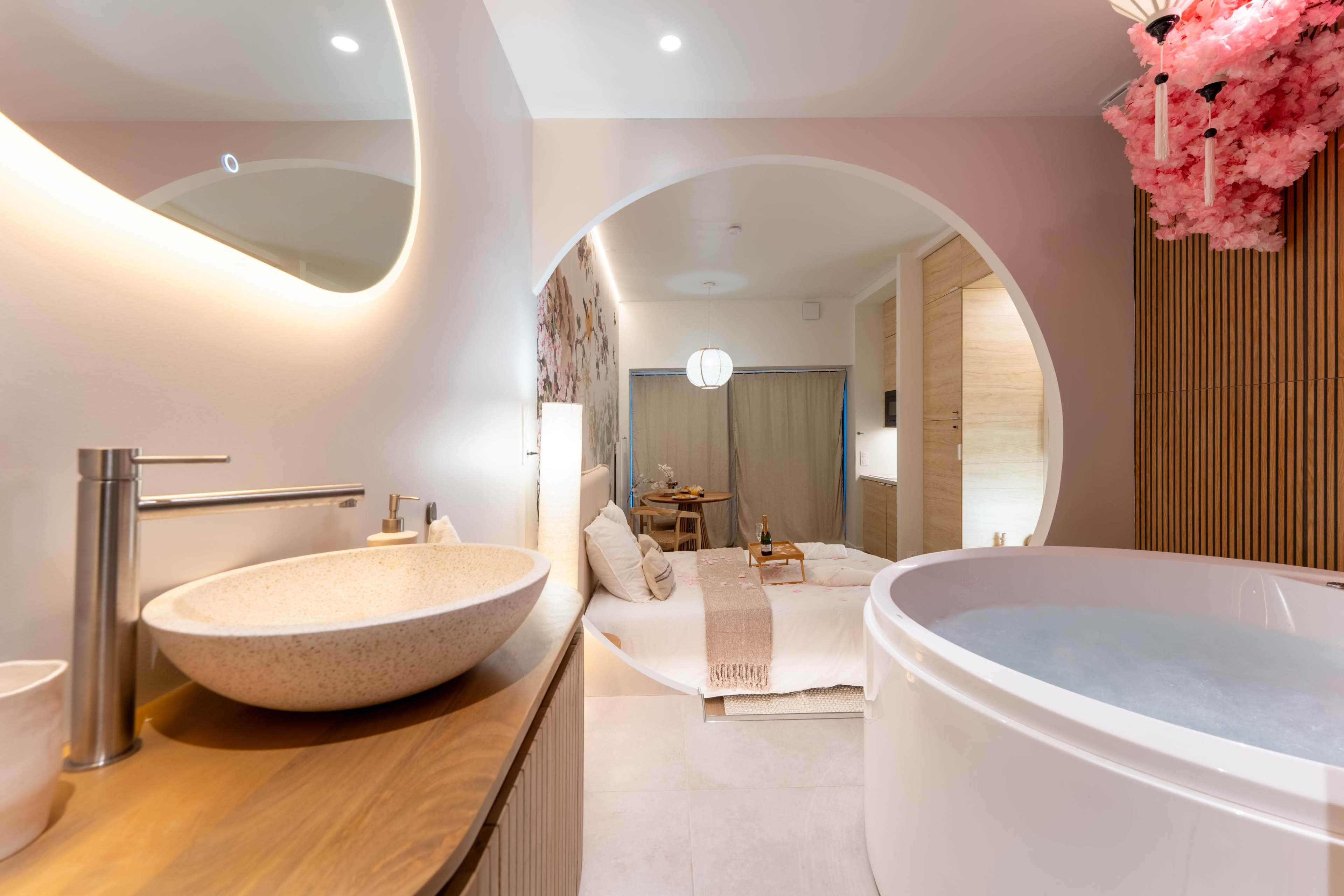 The image depicts a modern bathroom featuring a round soaking tub and a vessel sink, with a cozy bedroom area visible in the background.